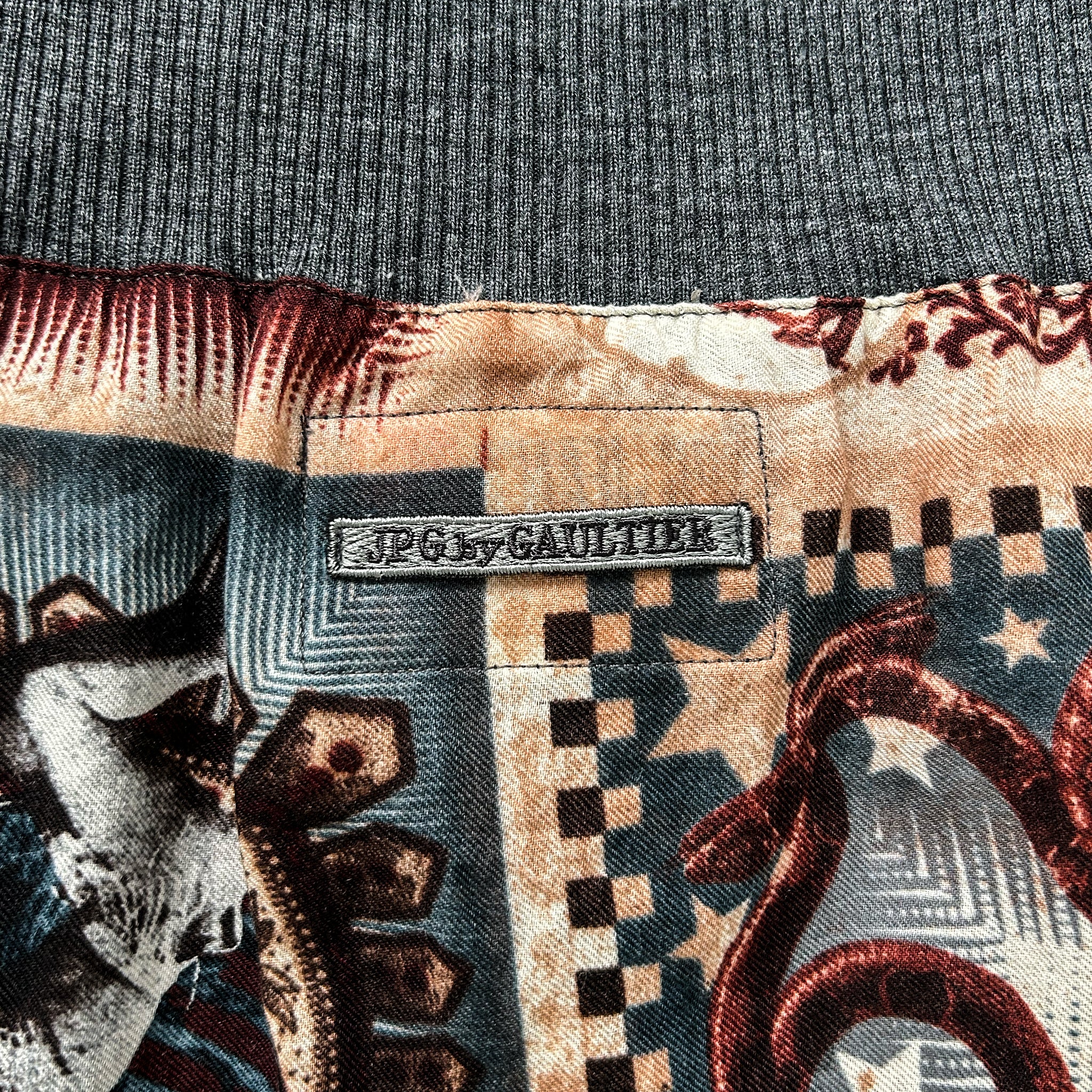 a close-up view of a fabric with a pattern featuring a snake and other geometric shapes. There is a metal tag or label attached to the fabric, which reads "J.P.G. by Gaultier".
