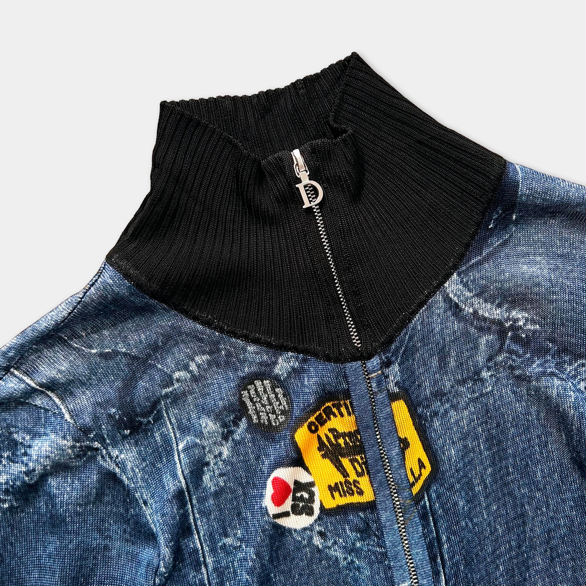 a close-up view of a blue denim jacket with a black zipper and a ribbed collar. The jacket has several patches and buttons attached to it, including a yellow patch with the text "MISS" and a black patch with the text "D".