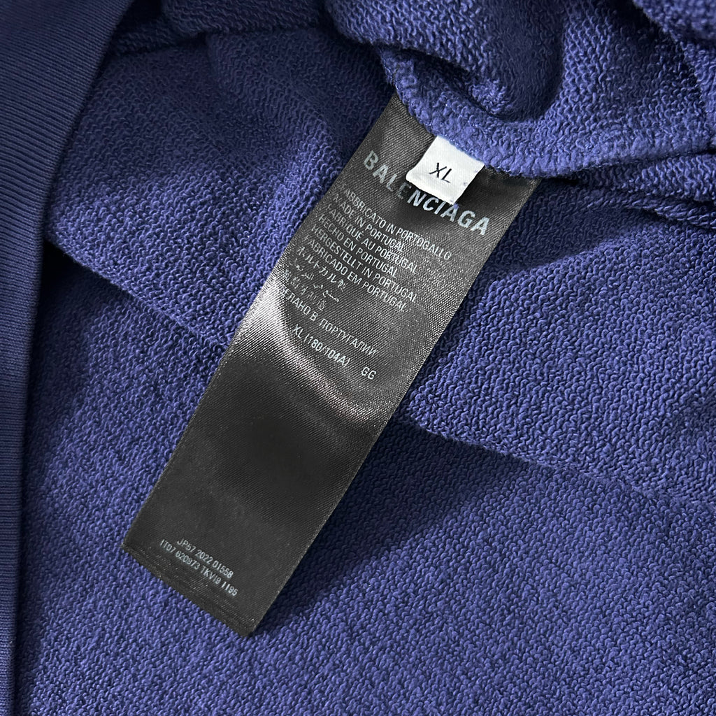 A close-up view of a dark blue fabric, possibly a sweater or garment, with a black label attached to it. The label appears to be a garment tag from a clothing brand, with text providing information about the product.