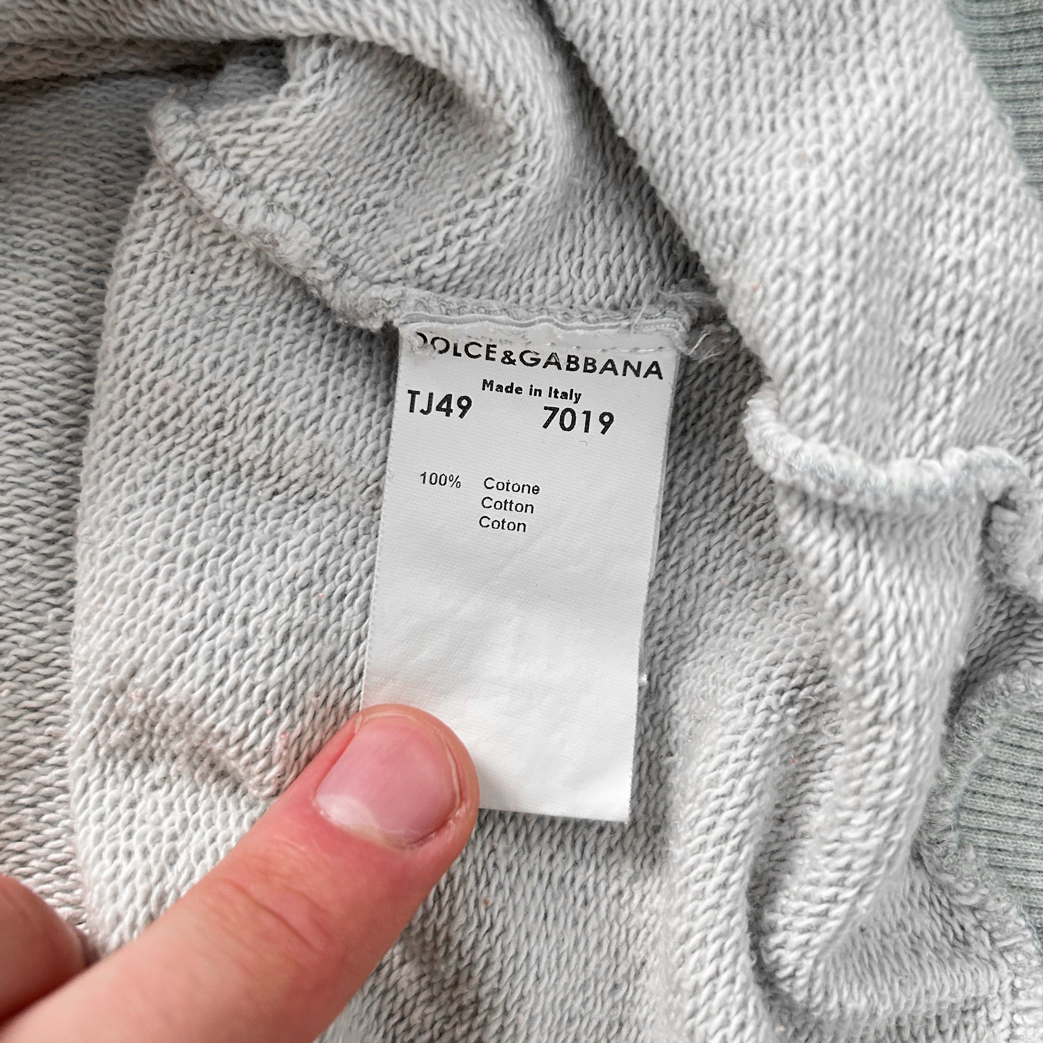 A close-up view of a white fabric, possibly a sweater or garment, with a label attached to it. The label appears to be from a brand like Dolce & Gabbana, indicating that the fabric is 100% cotton and made in Italy.