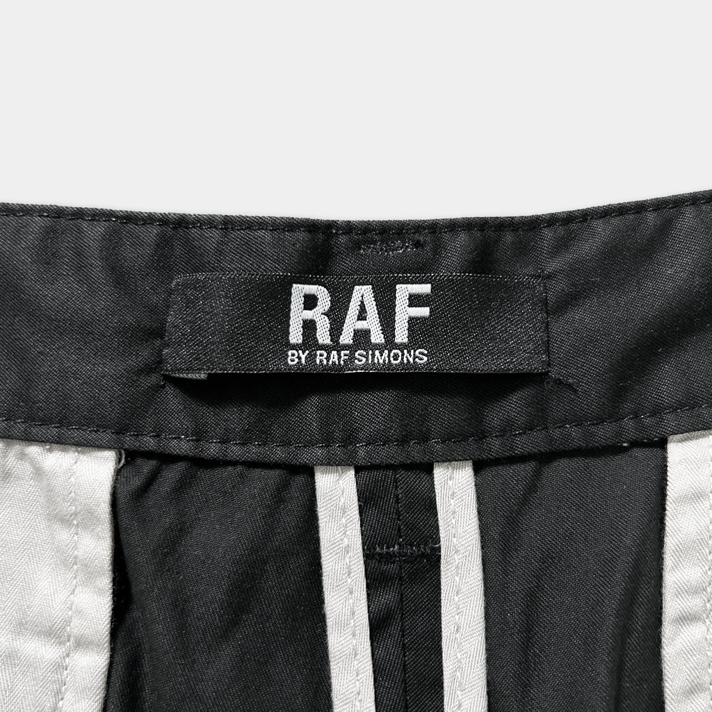 a close-up view of a black and white label on a pair of pants, which reads "RAF" and "BY RAF SIMONS".