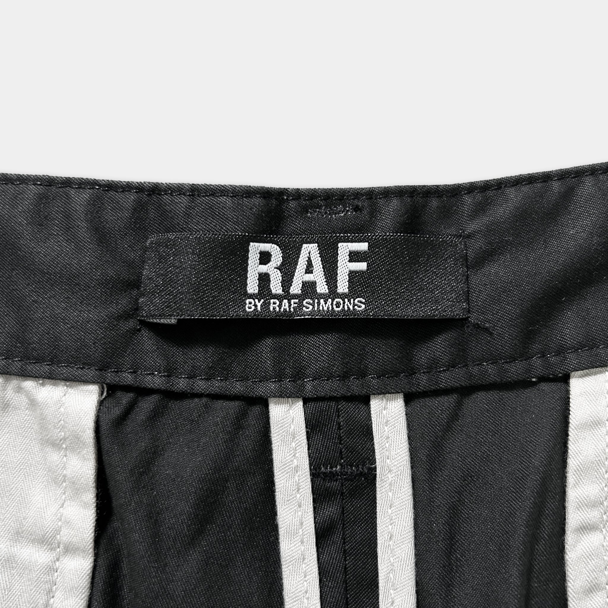 a close-up view of a black and white label on a pair of pants, which reads "RAF" and "BY RAF SIMONS".