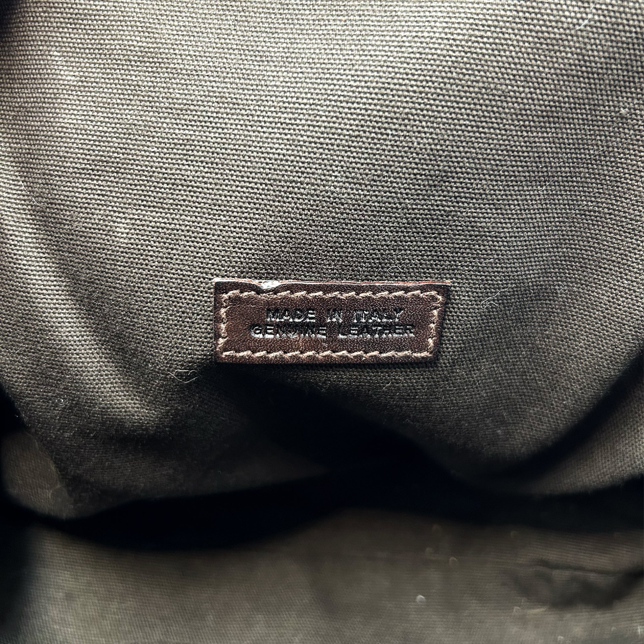 a close-up view of a fabric or leather material, with a small label or tag attached to it. The label appears to be made of leather and has text printed on it, indicating that it is genuine leather.