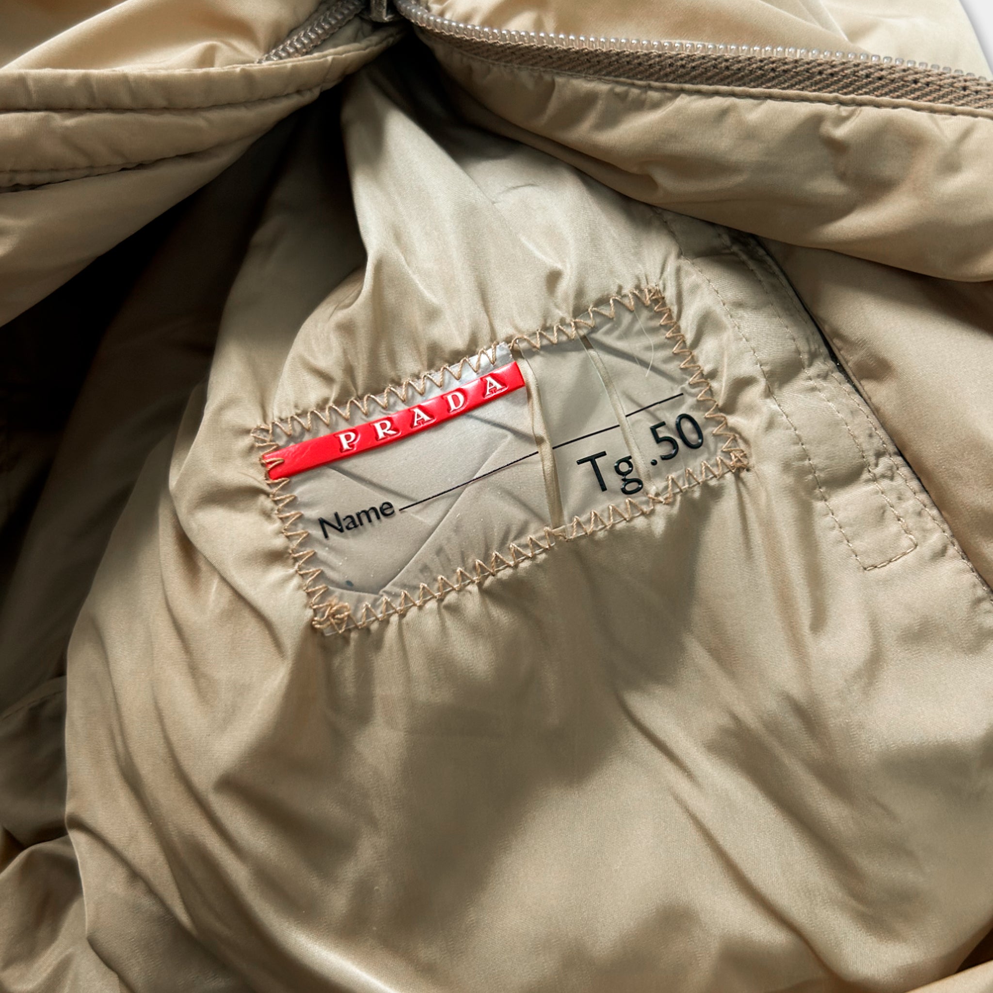 a close-up view of a beige fabric, possibly a jacket or sweater, with a red label attached to it. The label appears to be a name tag or a label with a price tag, indicating the brand and price of the item.