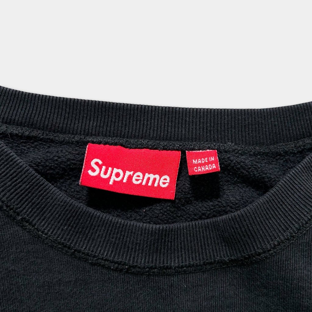 A close-up view of a black sweater or shirt with a red Supreme logo and a red tag indicating that it is made in Canada.