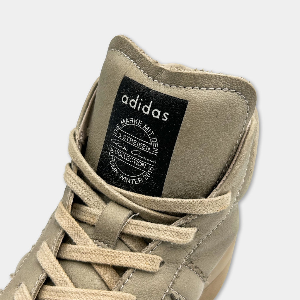 A close-up view of a tan Adidas sneaker, showing the brand's logo and a black label with text.