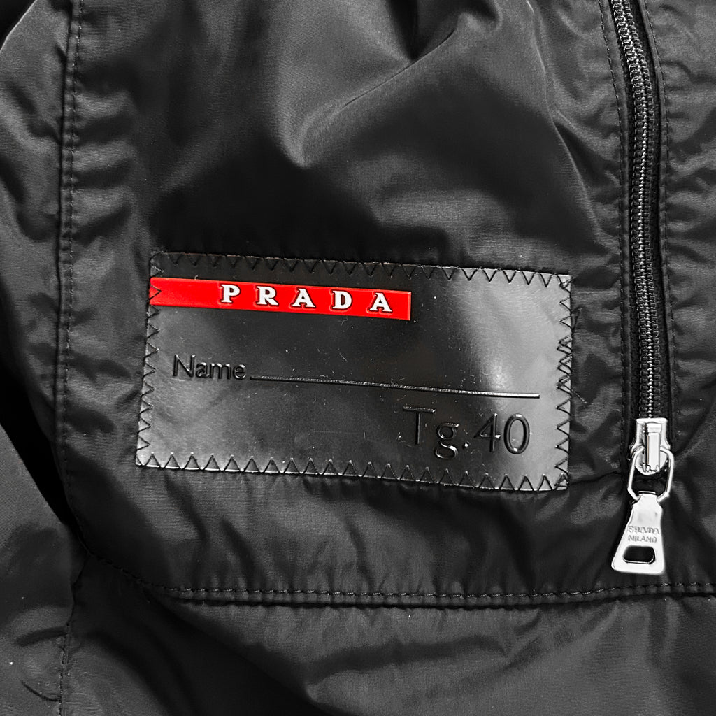 a close-up view of a black jacket with a red and white label on the front. The label appears to be a name tag or patch, and it has the word "PRADA" printed on it.