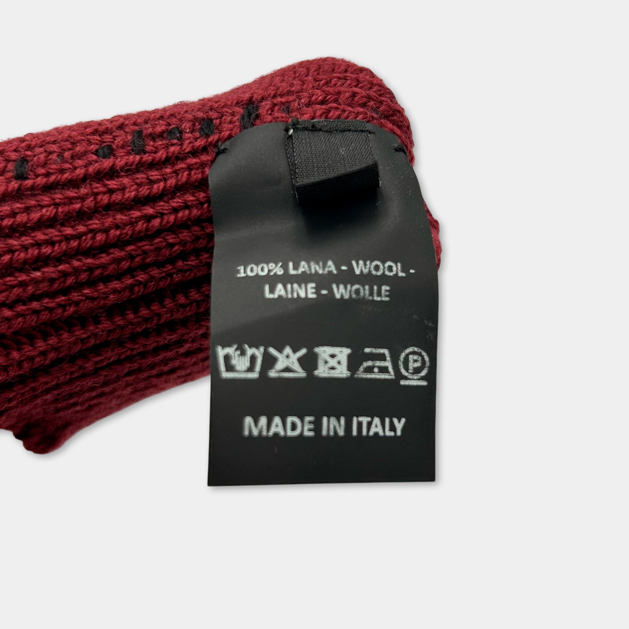 A close-up image of a red knitted scarf with a black tag attached, displaying information about the scarf's material and origin.