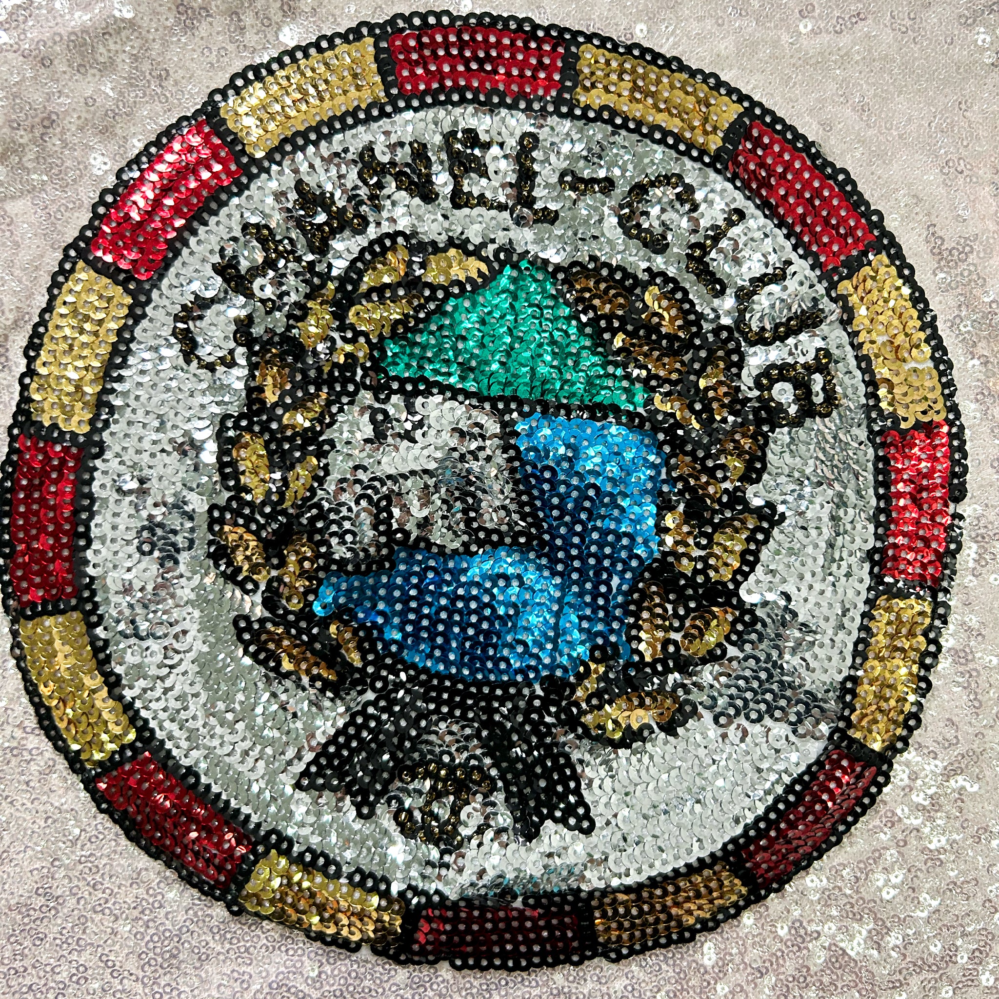 The image is a circular emblem or badge with a vibrant, colorful design. The emblem is composed of numerous small sequin beads arranged in a circular pattern, creating a visually striking and intricate design.