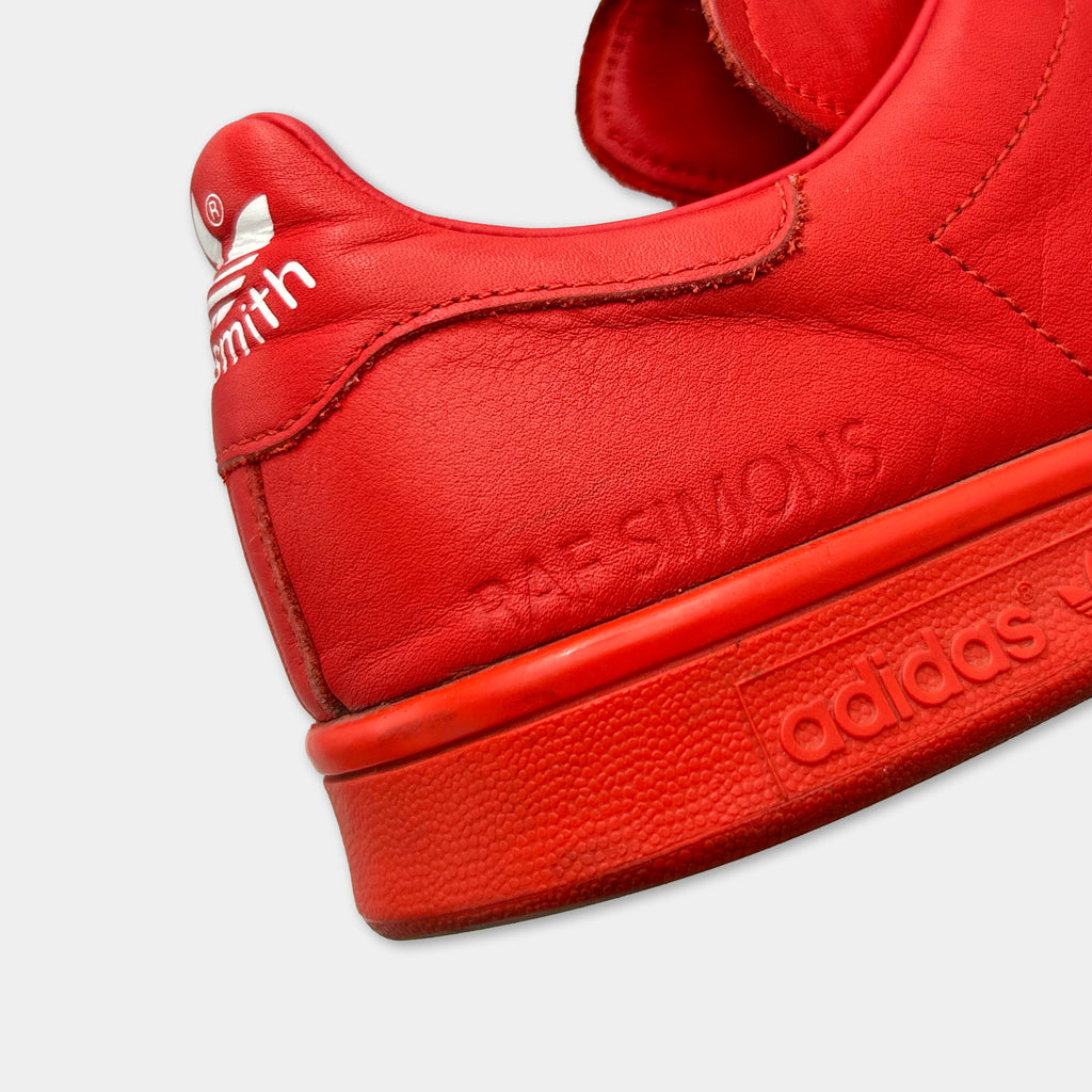 A close-up of a red Adidas sneaker with the brand name and the name of the designer, Raf Simons, visible on the side.