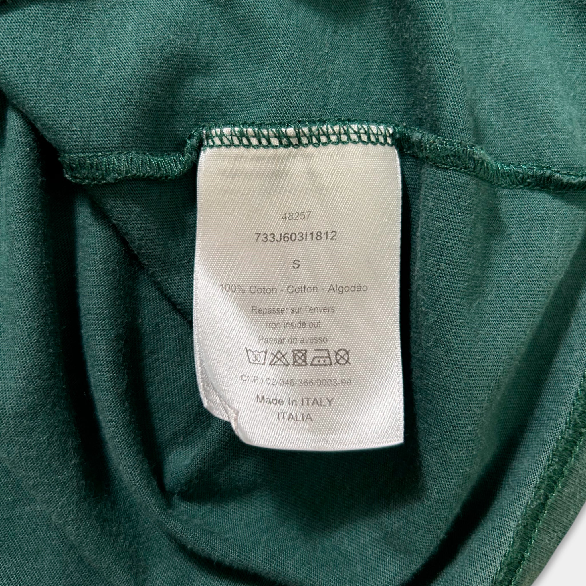 a close-up view of a green fabric, possibly a t-shirt or garment, with a white label attached to it. The label contains text and symbols, likely providing information about the product, such as the brand, material, and care instructions.