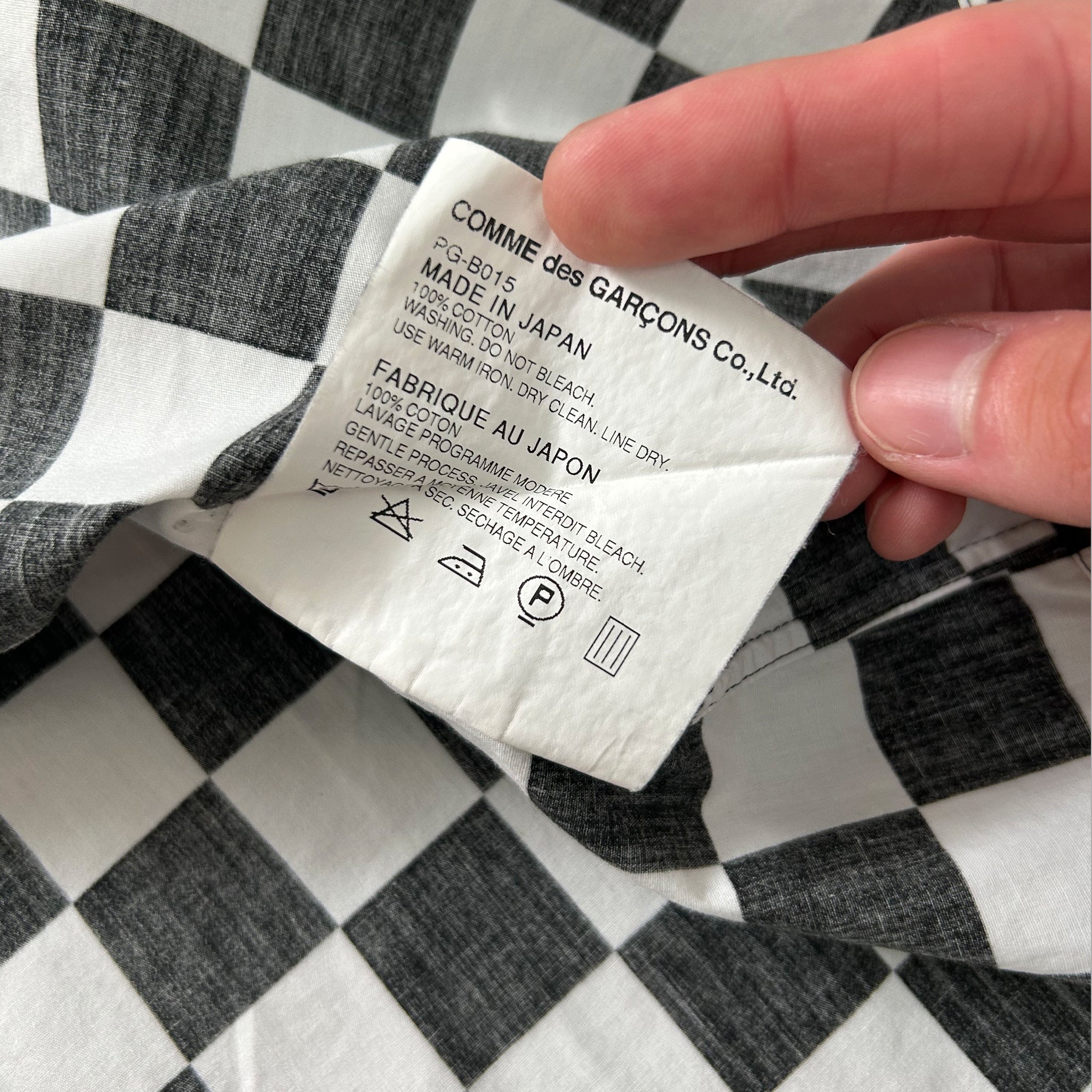 A hand is holding a label with text on it, which appears to be a label from a garment. The label is placed on a fabric with a checkered pattern.