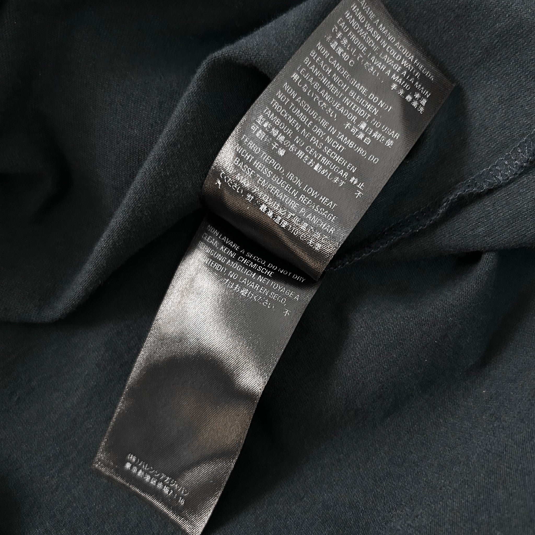 a close-up view of a black fabric, possibly a t-shirt or garment, with a label attached to it. The label appears to contain text in a foreign language, which is not transcribed due to the language barrier. The image does not contain any other objects or text, and the focus is solely on the label and the fabric.