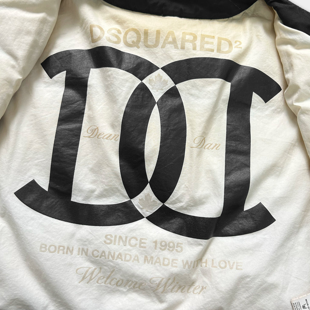 a close-up view of a white jacket with a large black logo on the back. The logo appears to be a stylized "D" or "C" with a leaf design, and it is accompanied by text that reads "DQSQUARED2" and "Since 1995", "Born in Canada Made with Love", and "Welcome Winter".