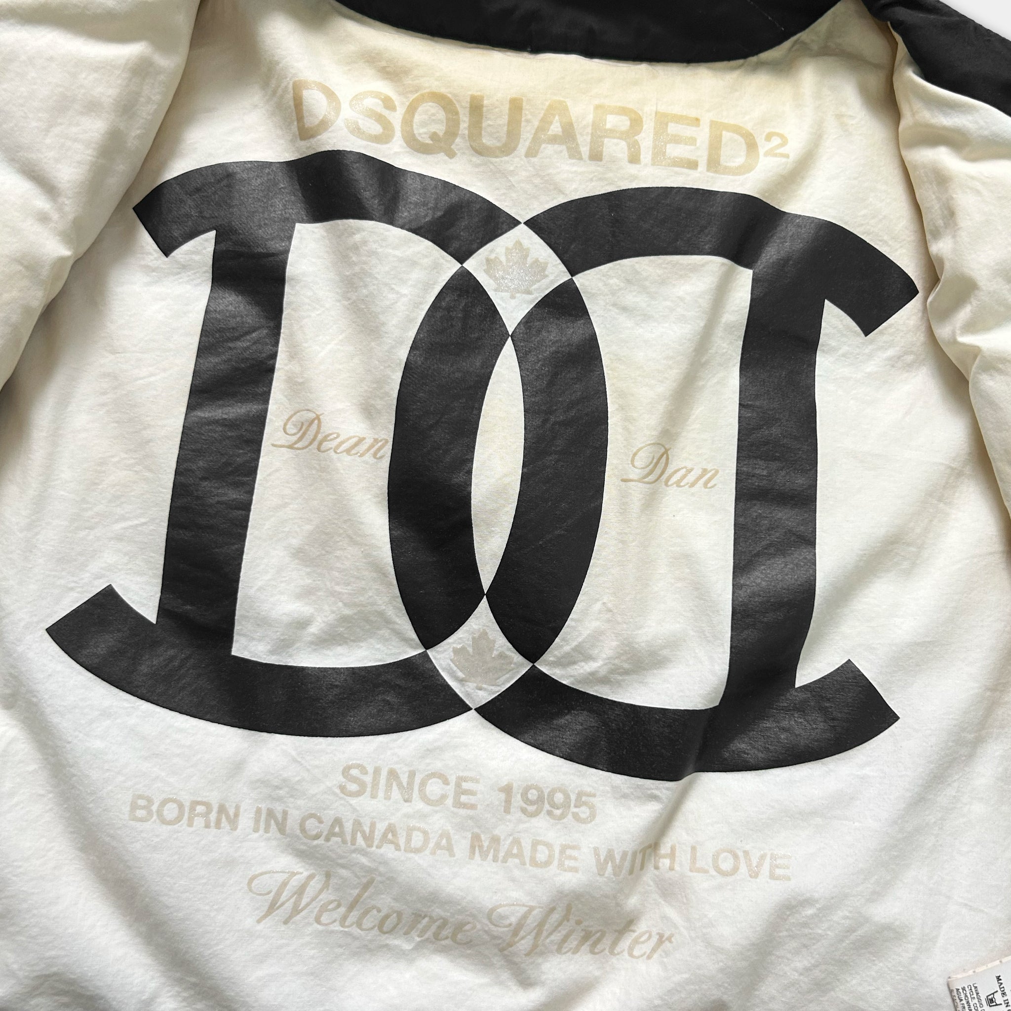 a close-up view of a white jacket with a large black logo on the back. The logo appears to be a stylized "D" or "C" with a leaf design, and it is accompanied by text that reads "DQSQUARED2" and "Since 1995", "Born in Canada Made with Love", and "Welcome Winter".
