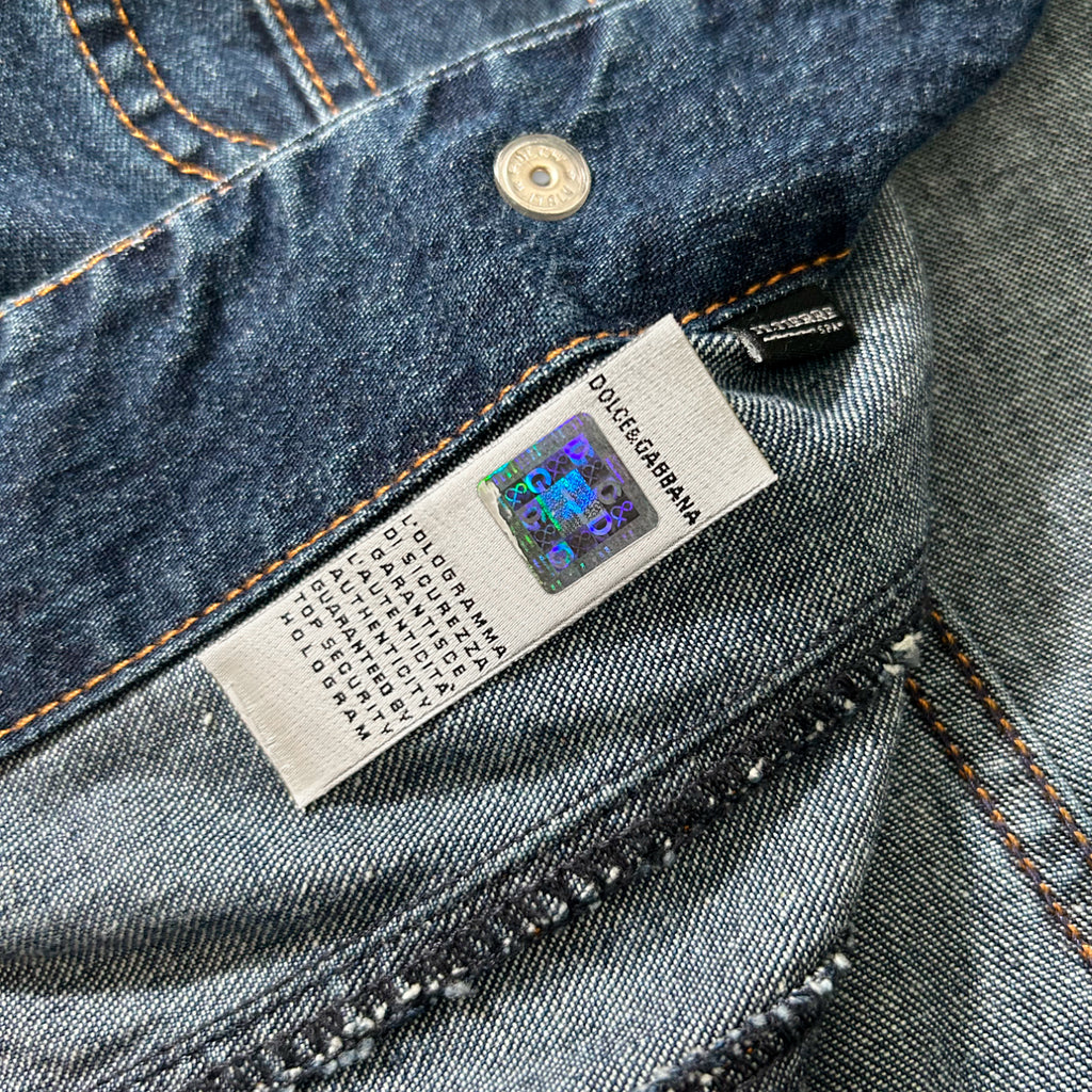 a close-up view of a denim fabric, possibly a pair of jeans, with a label attached to it. The label appears to be a product tag from a brand like Dolce & Gabbana, indicating the brand and possibly the product's details.