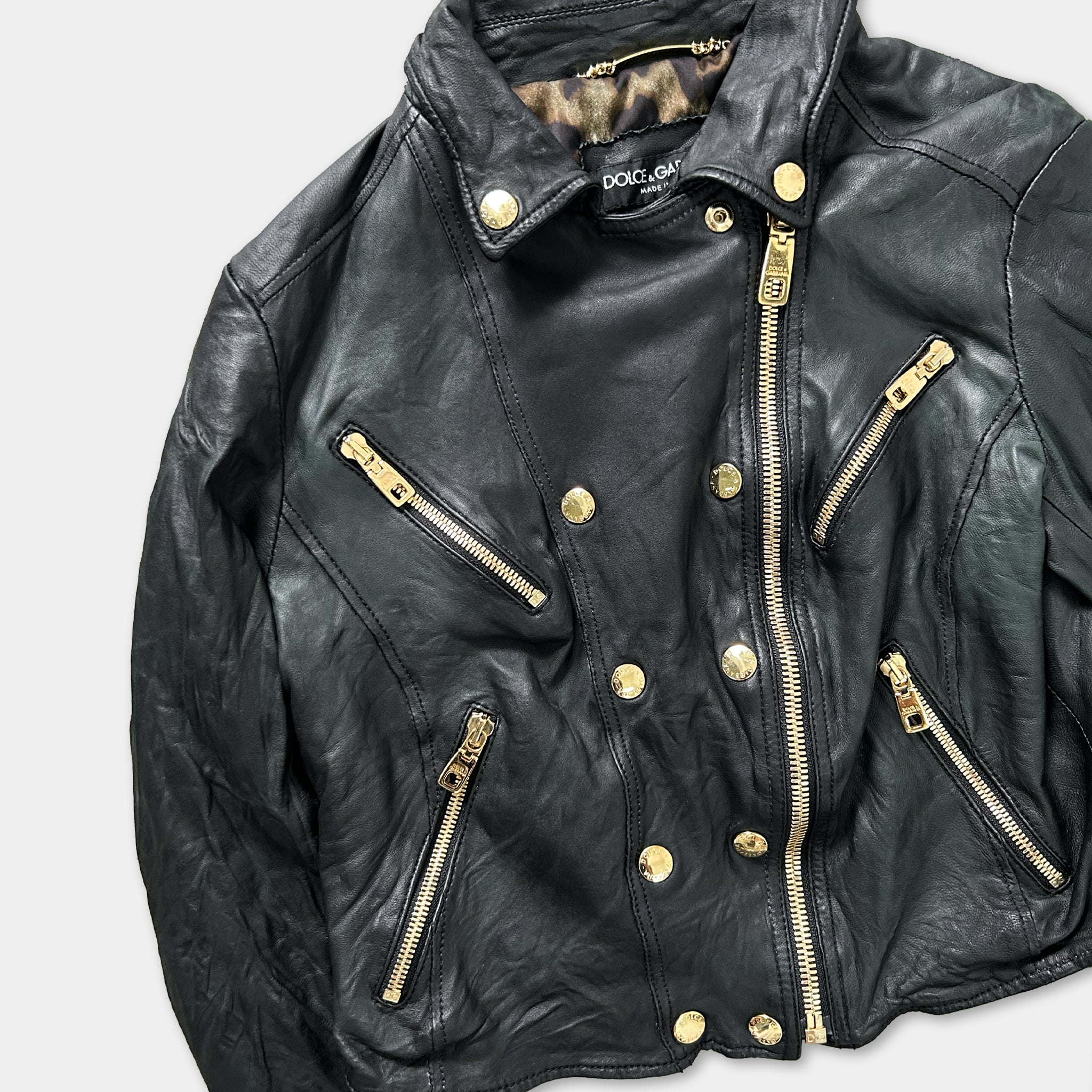 A black leather jacket with gold zippers and buttons, featuring a collar and multiple pockets.