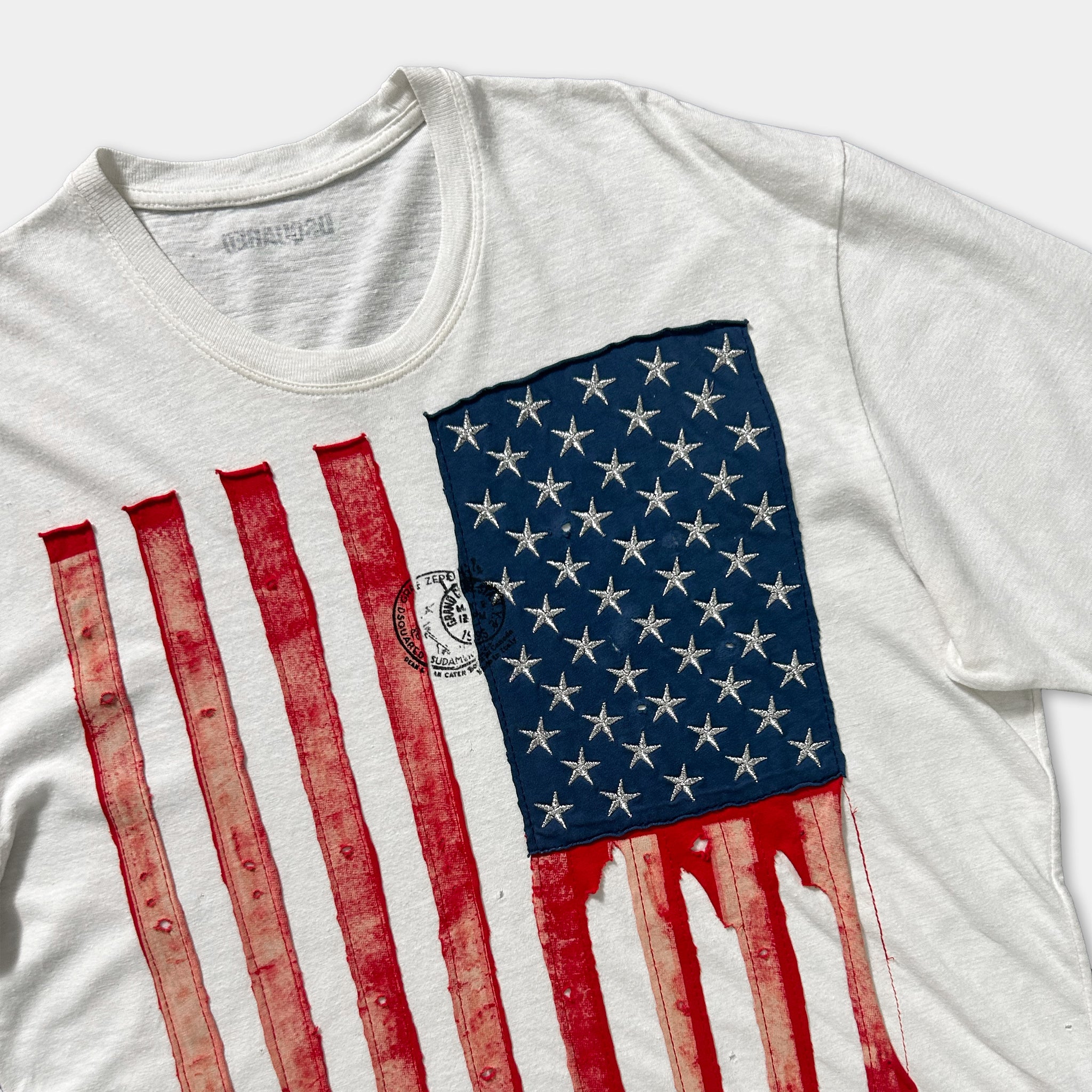 a white t-shirt with a prominent American flag design, featuring red and white stripes and a blue patch with white stars.