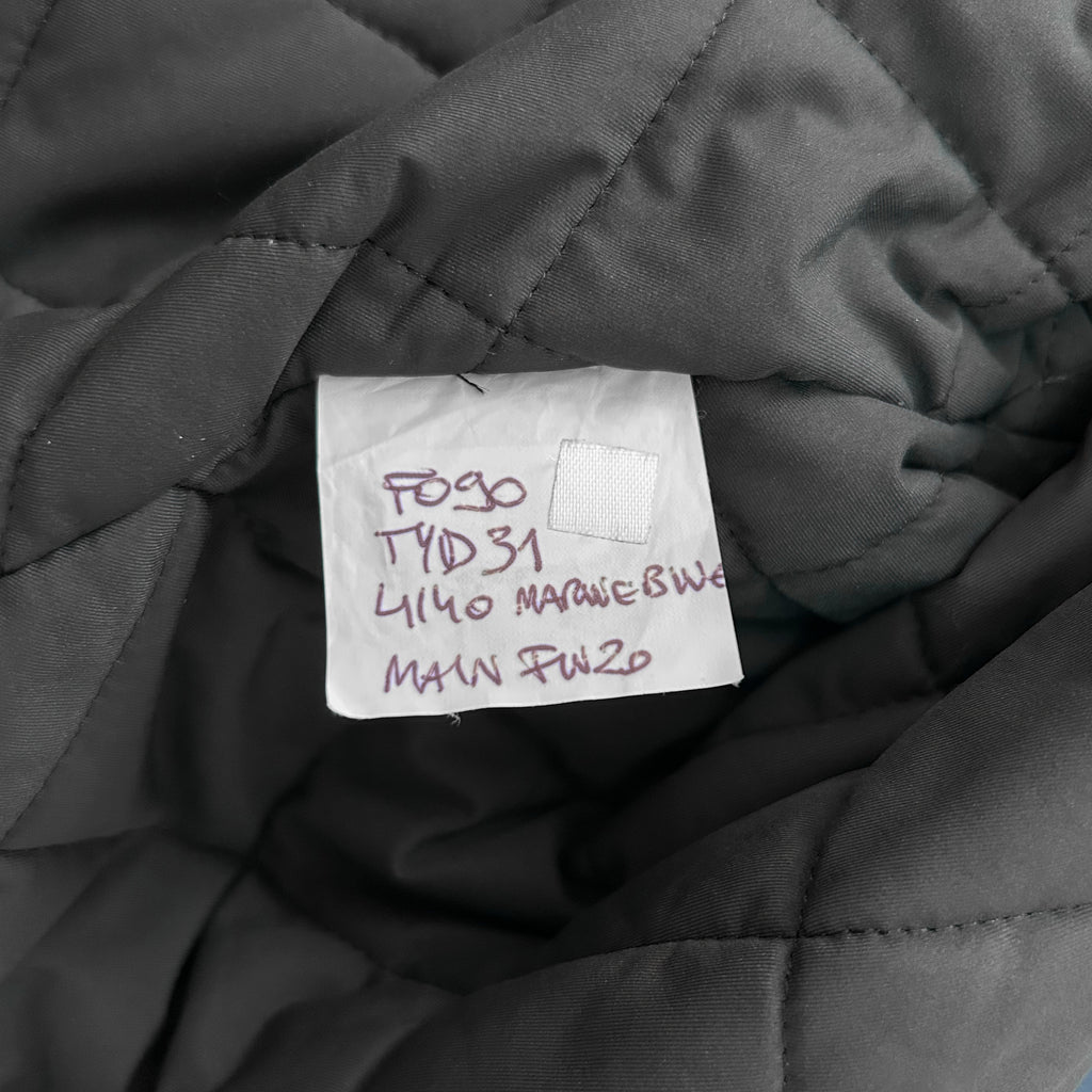 a close-up view of a gray fabric, possibly a blanket or quilt, with a handwritten label attached to it. The label appears to be a price tag or inventory label, with handwritten text that is not fully legible.