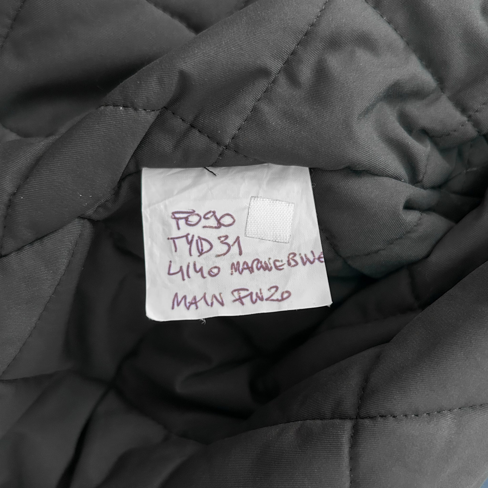 a close-up view of a gray fabric, possibly a blanket or quilt, with a handwritten label attached to it. The label appears to be a price tag or inventory label, with handwritten text that is not fully legible.