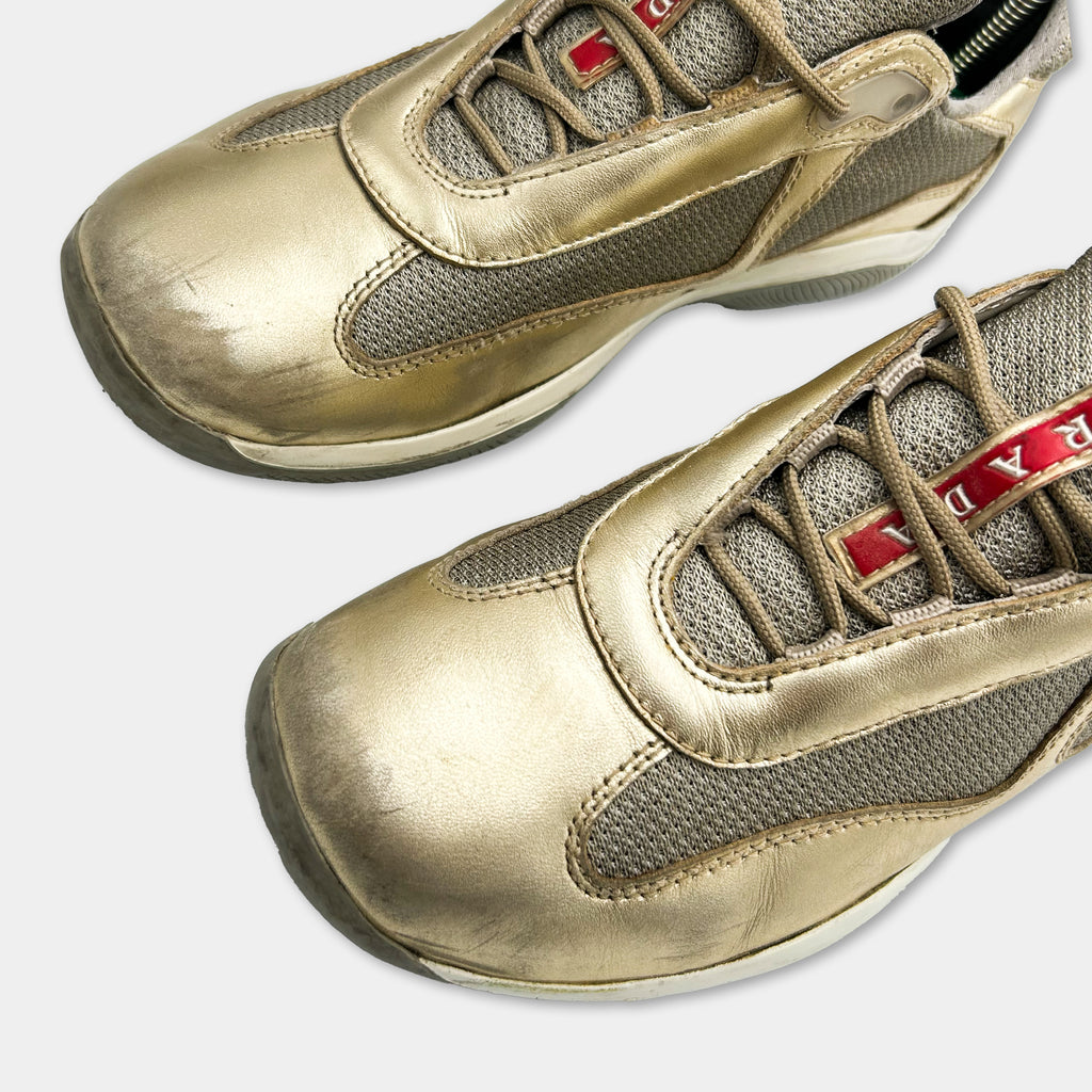A close-up view of a pair of gold-colored athletic shoes with a mesh upper and a red lacing system.