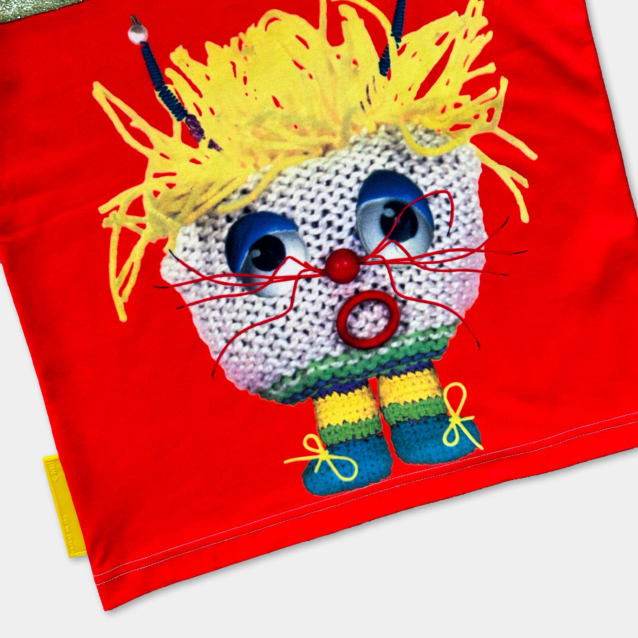 A red fabric with a whimsical, hand-knitted character design featuring a yellow hat, red nose, and blue and green striped socks.