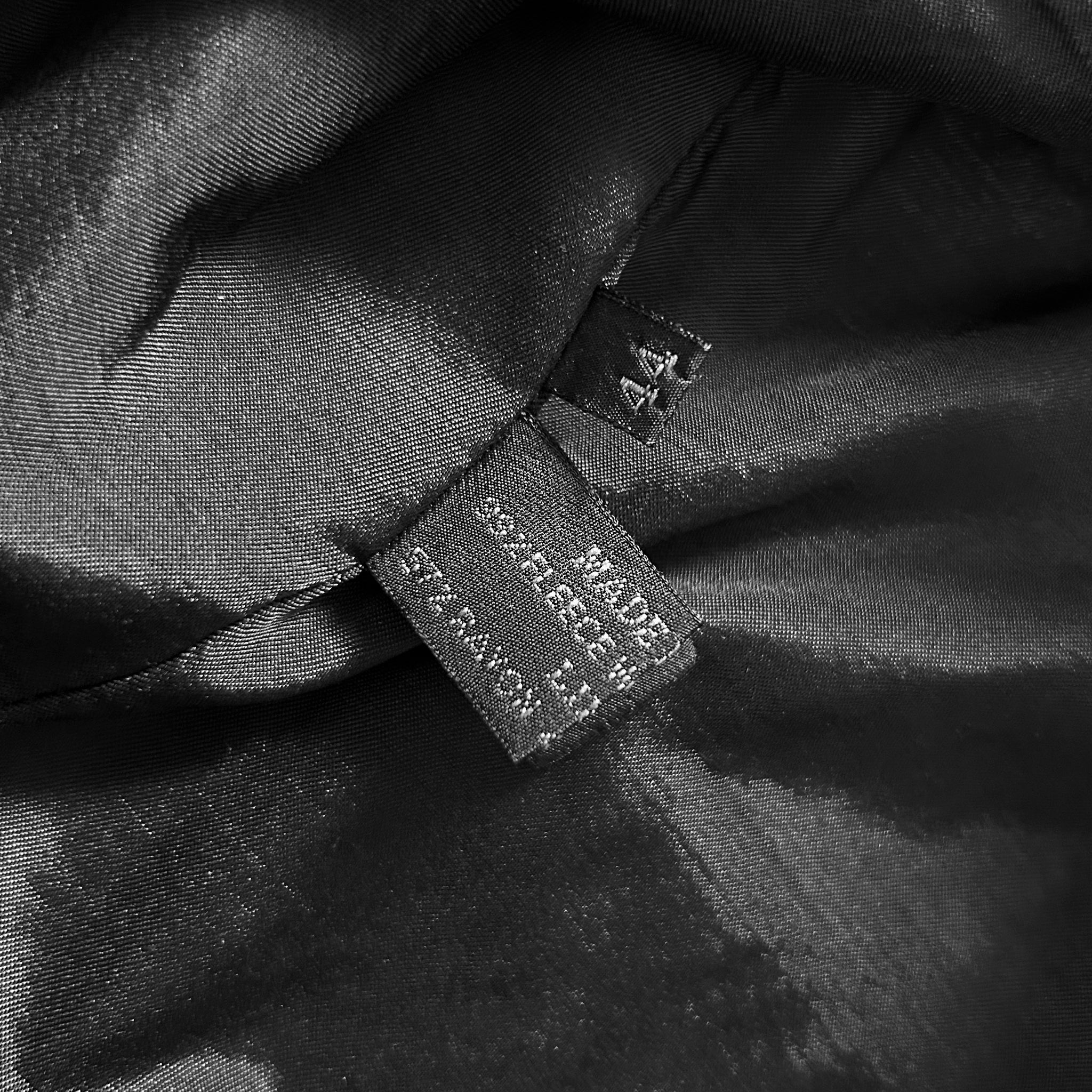 a close-up view of a black fabric, possibly a piece of clothing or a textile. There is a small label or tag attached to the fabric, which contains text that is partially obscured. The visible text appears to be related to the material or the product, but it is not clear enough to transcribe accurately.