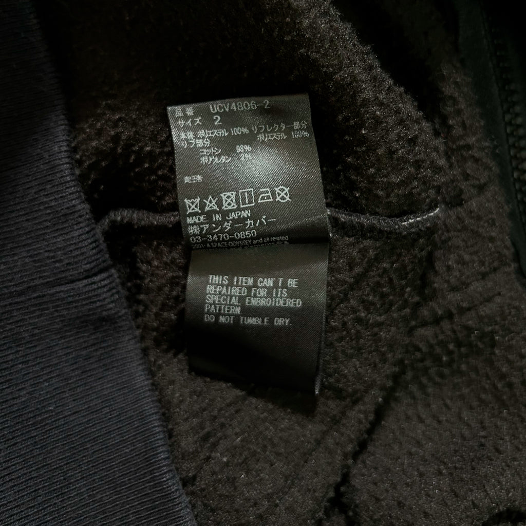 a close-up view of a black fabric, possibly a sweater or a jacket, with a label attached to it. The label contains text in Japanese and English, providing information about the product.
