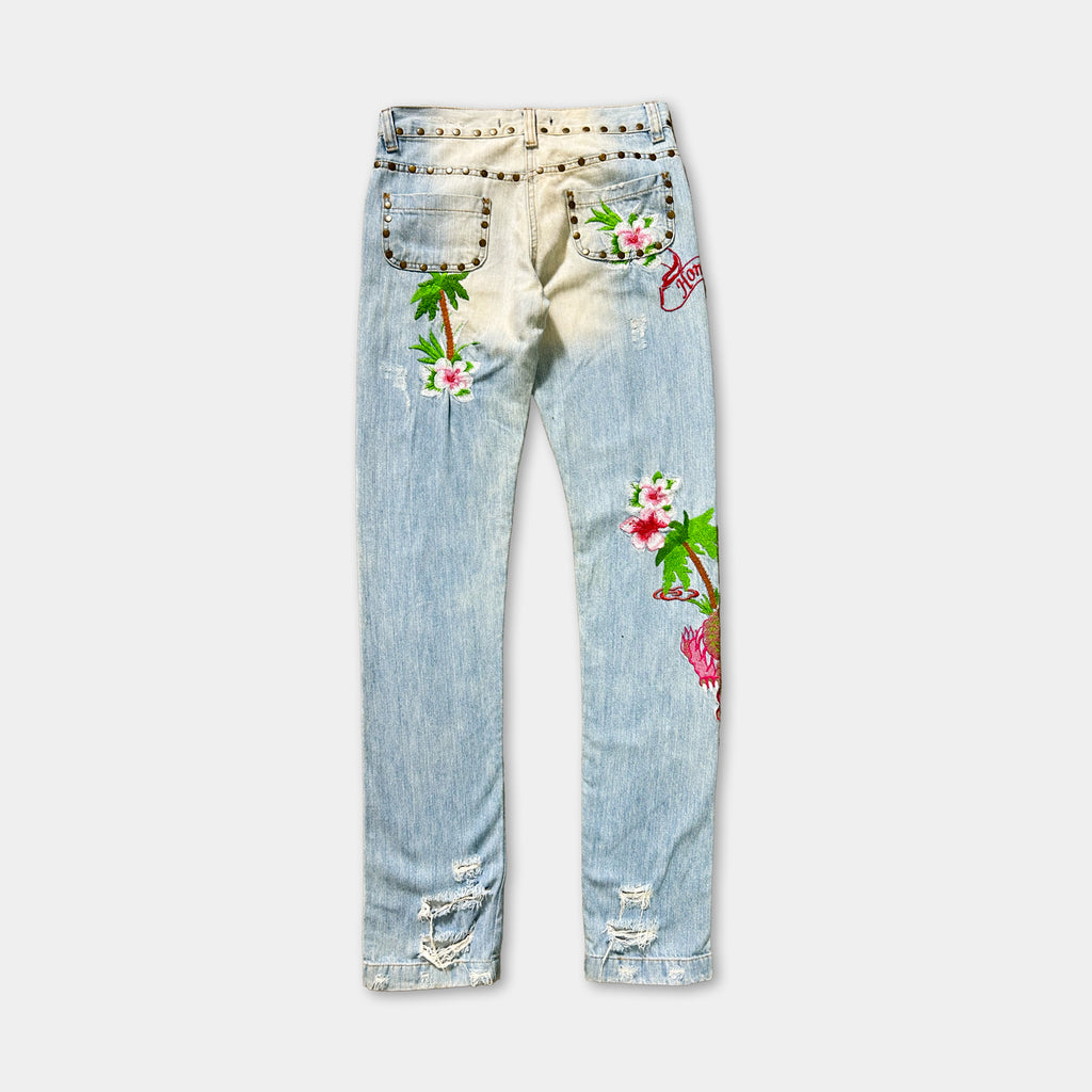A pair of light blue denim jeans with floral and palm tree designs on the back pockets and legs.