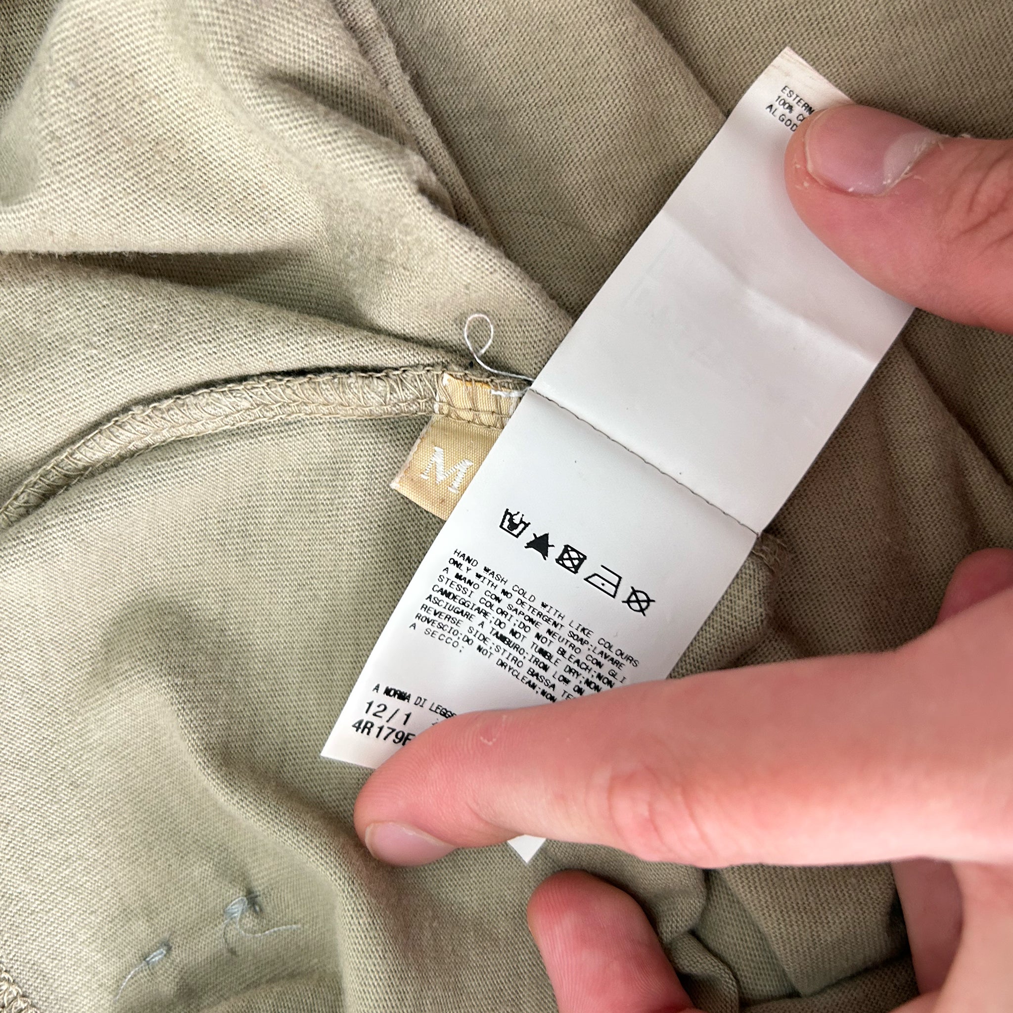 A person's hand is holding a white label with black text, which appears to be a garment label.