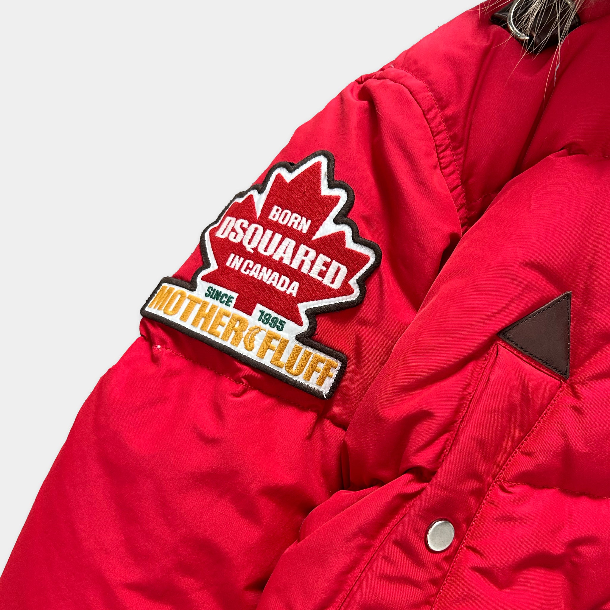A red puffy jacket with a red and white patch on the left arm that says "Born D'SQUARED IN Canada" and "Motherfluff" on the right arm.