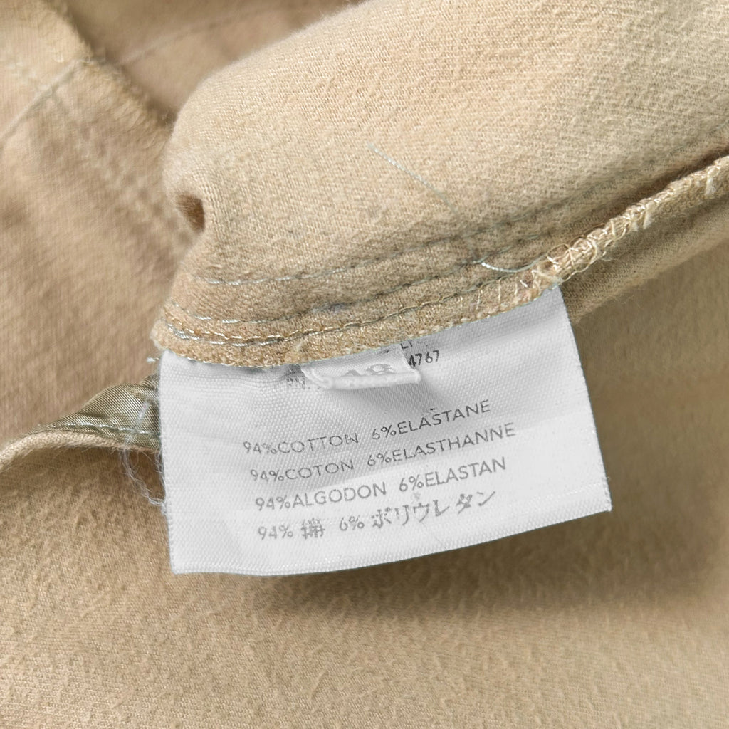 A close-up view of a beige fabric, possibly a piece of clothing, with a label attached to it. The label contains text in Swedish, indicating the fabric as 94% cotton, 6% elastane, and 94% algodon.
