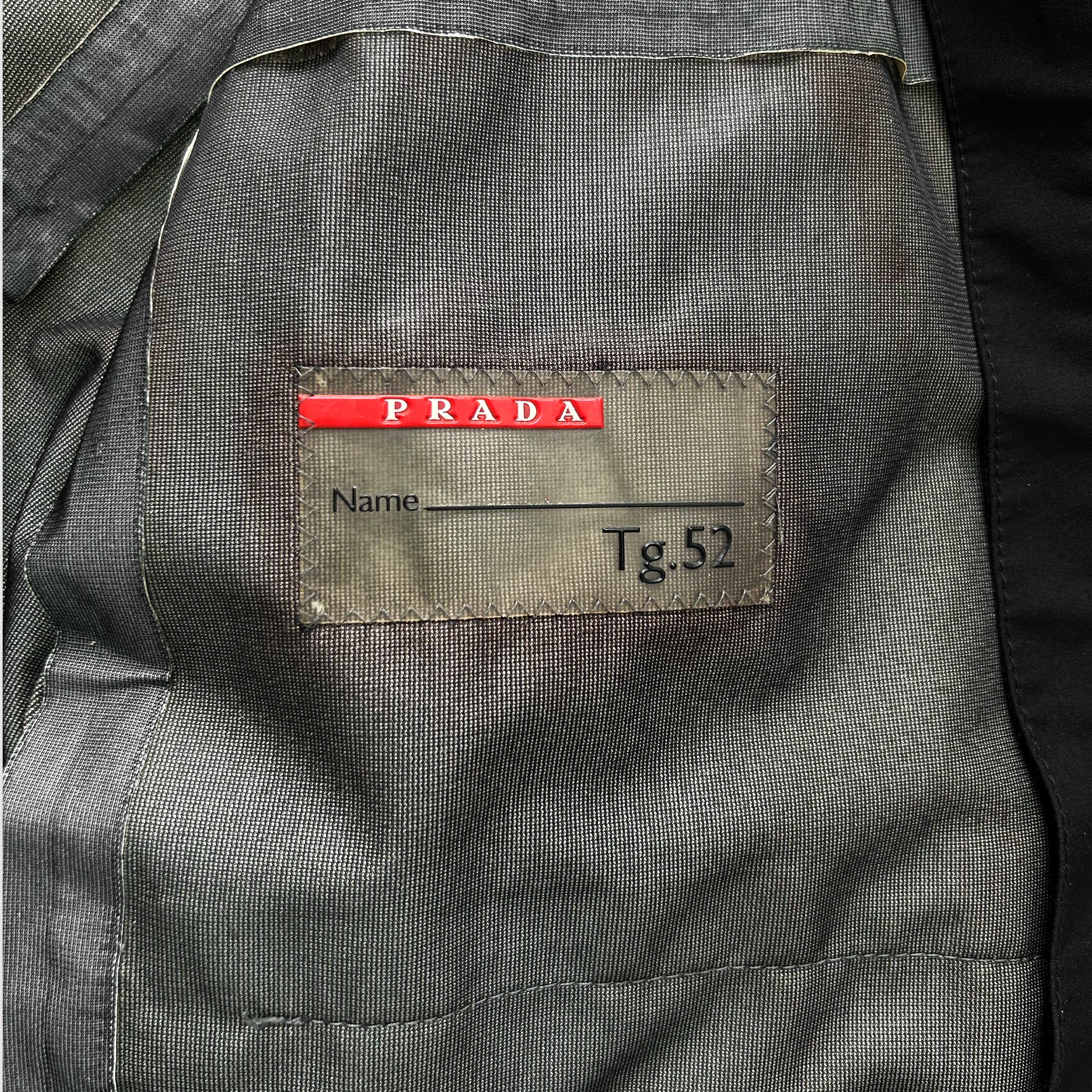 a gray fabric with a red label on it, which has the word "PRADA" printed on it. The label also has the name "Name" and the number "Tg52" printed on it.