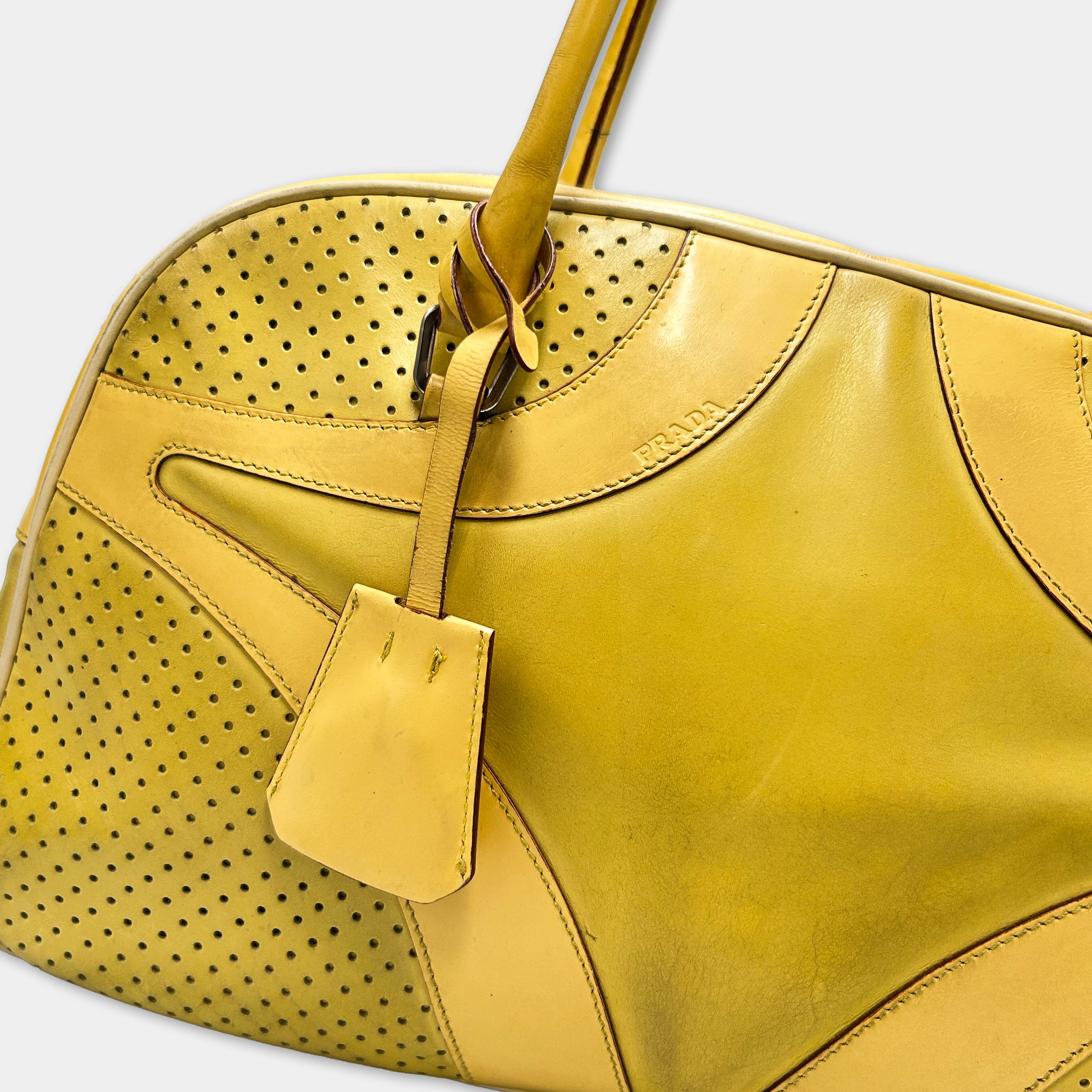 A close-up image of a yellow handbag with a perforated pattern and a small yellow tag attached to it.