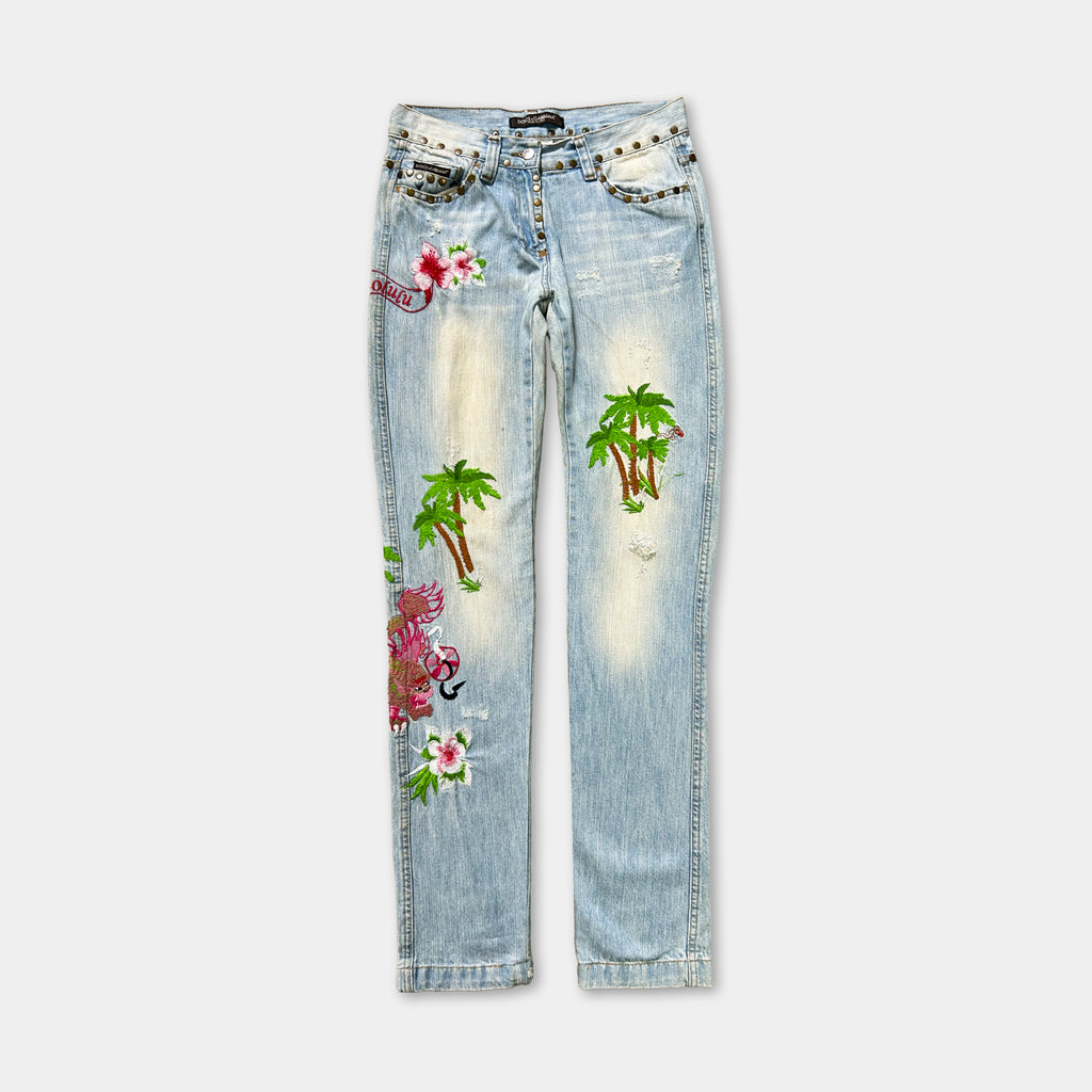 A pair of light blue denim jeans with various colorful and intricate embroidered designs, including palm trees, flowers, and other patterns.