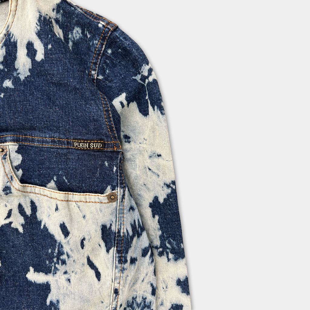 A close-up view of a denim jacket with a blue and white tie-dye pattern, featuring a pocket and a label on the front.