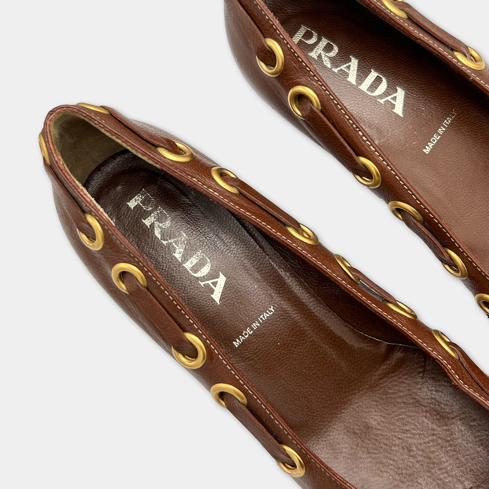 A pair of brown leather shoes with gold hardware, featuring the brand name "PRADA" printed on the side.