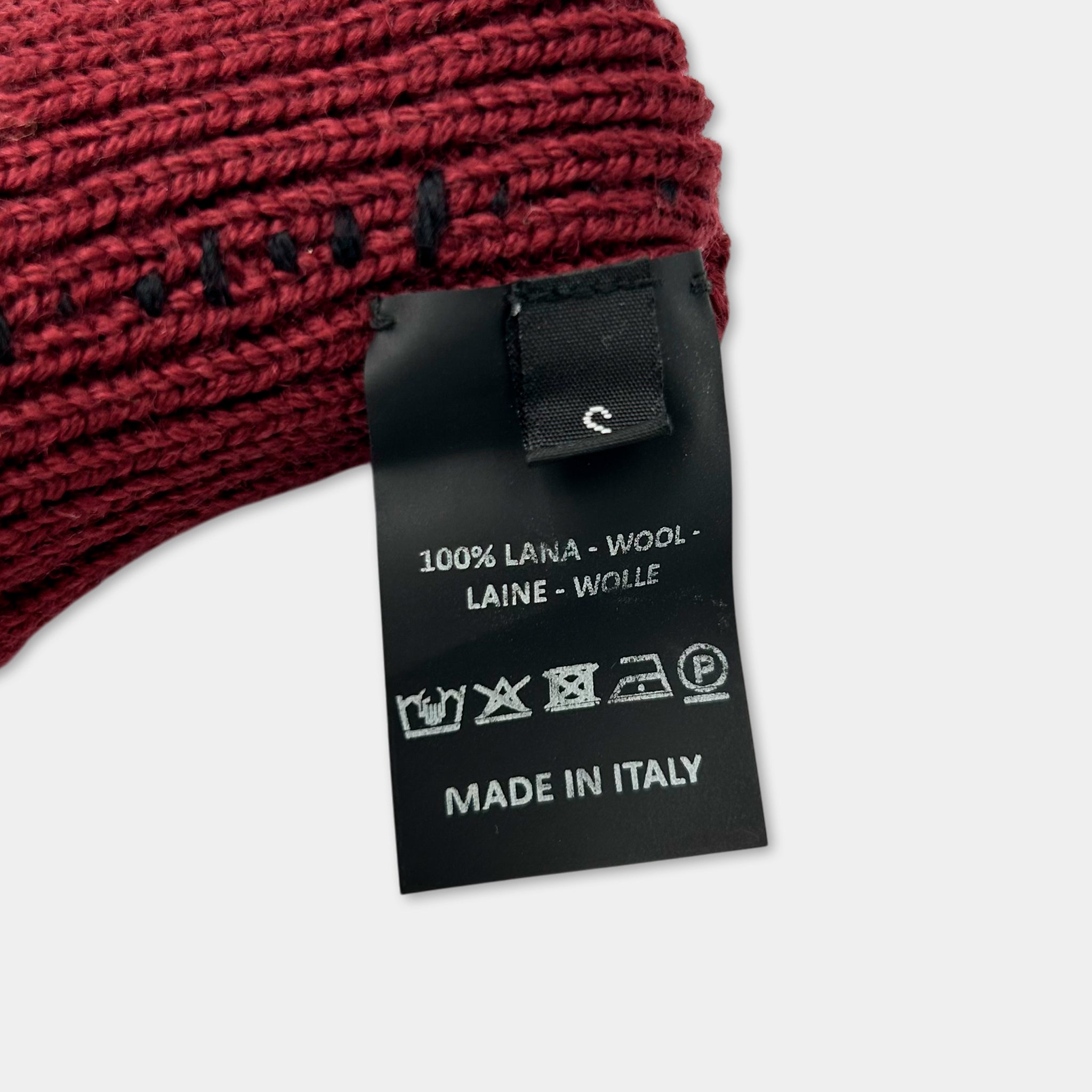 A close-up image of a red knitted hat with a black tag attached to it, displaying information about the product.