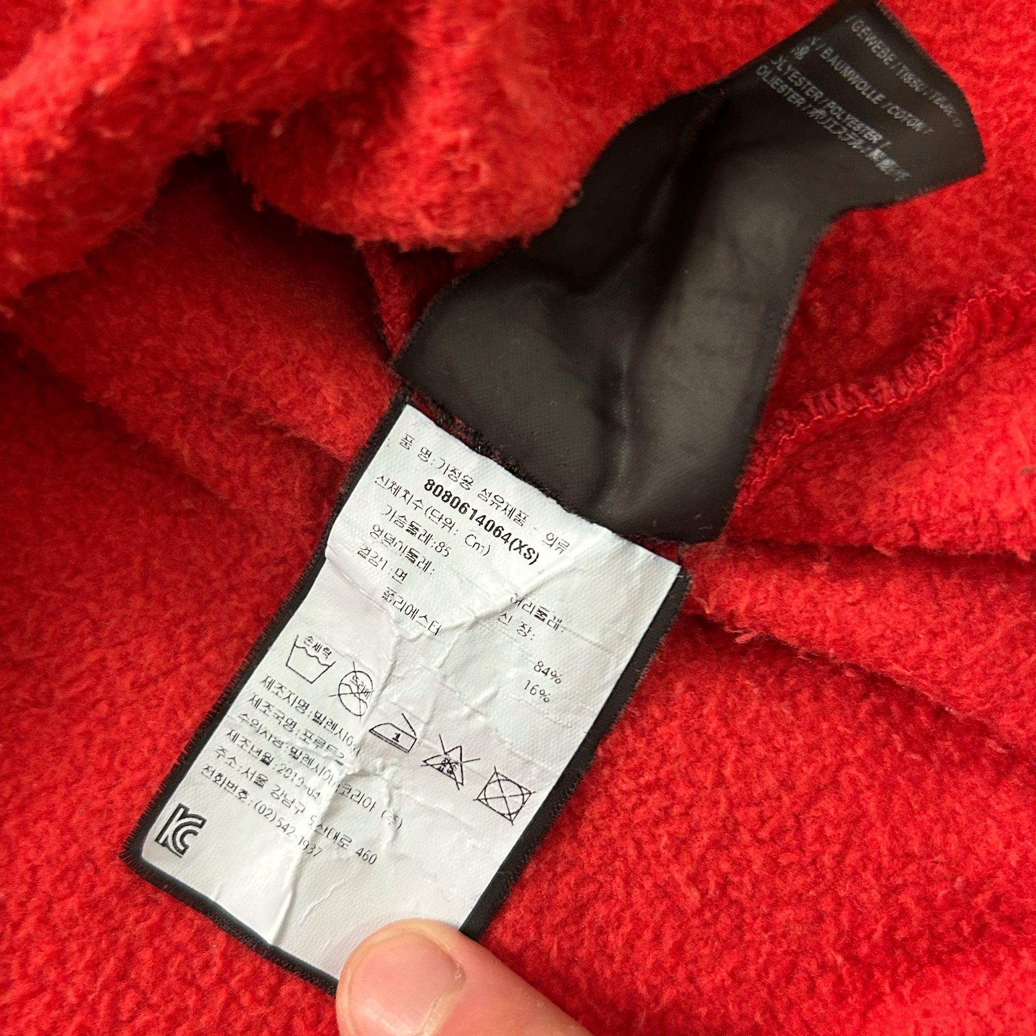 a close-up view of a red fabric, possibly a sweater or a blanket, with a black label attached to it. The label appears to contain text and symbols, but the image does not contain any text that can be transcribed due to its small size and the fact that it is a close-up view.