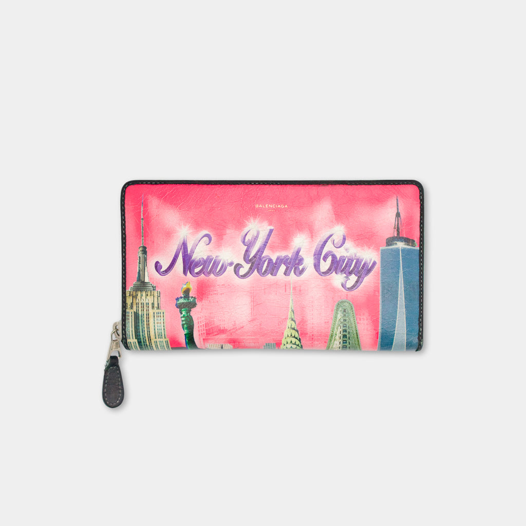 A pink wallet or card holder with the text "New York City" printed on it, featuring illustrations of iconic New York City landmarks such as the Empire State Building and the Statue of Liberty.