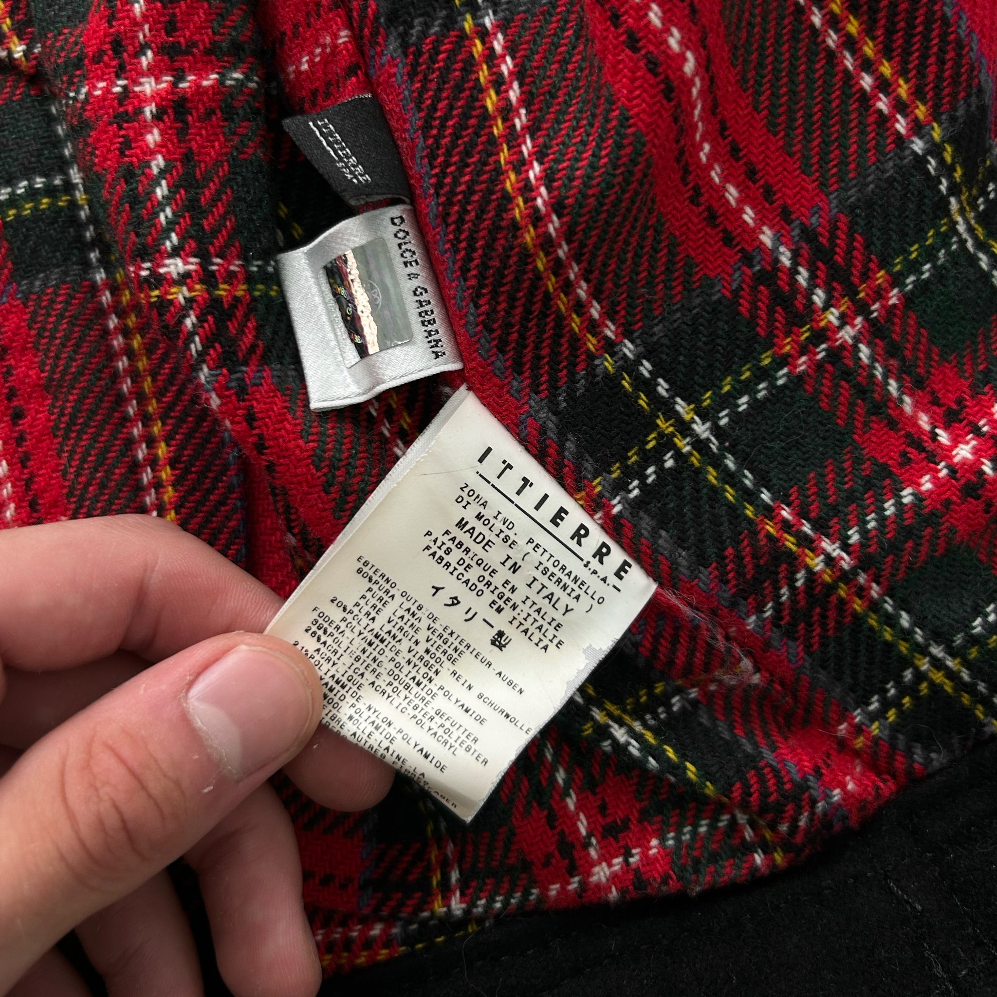 A hand is holding a label with text on it, which appears to be a label from a garment. The label is placed on a red and black plaid fabric.