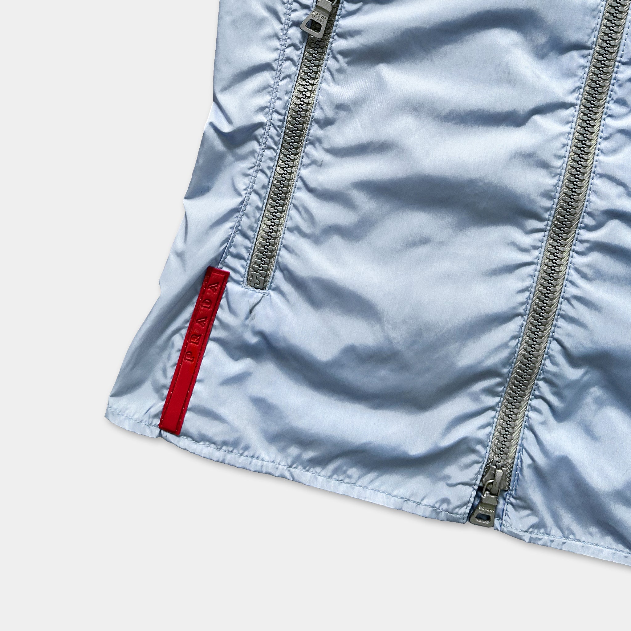 A close-up view of a light blue jacket with a red zipper pull and a red logo on the left side.