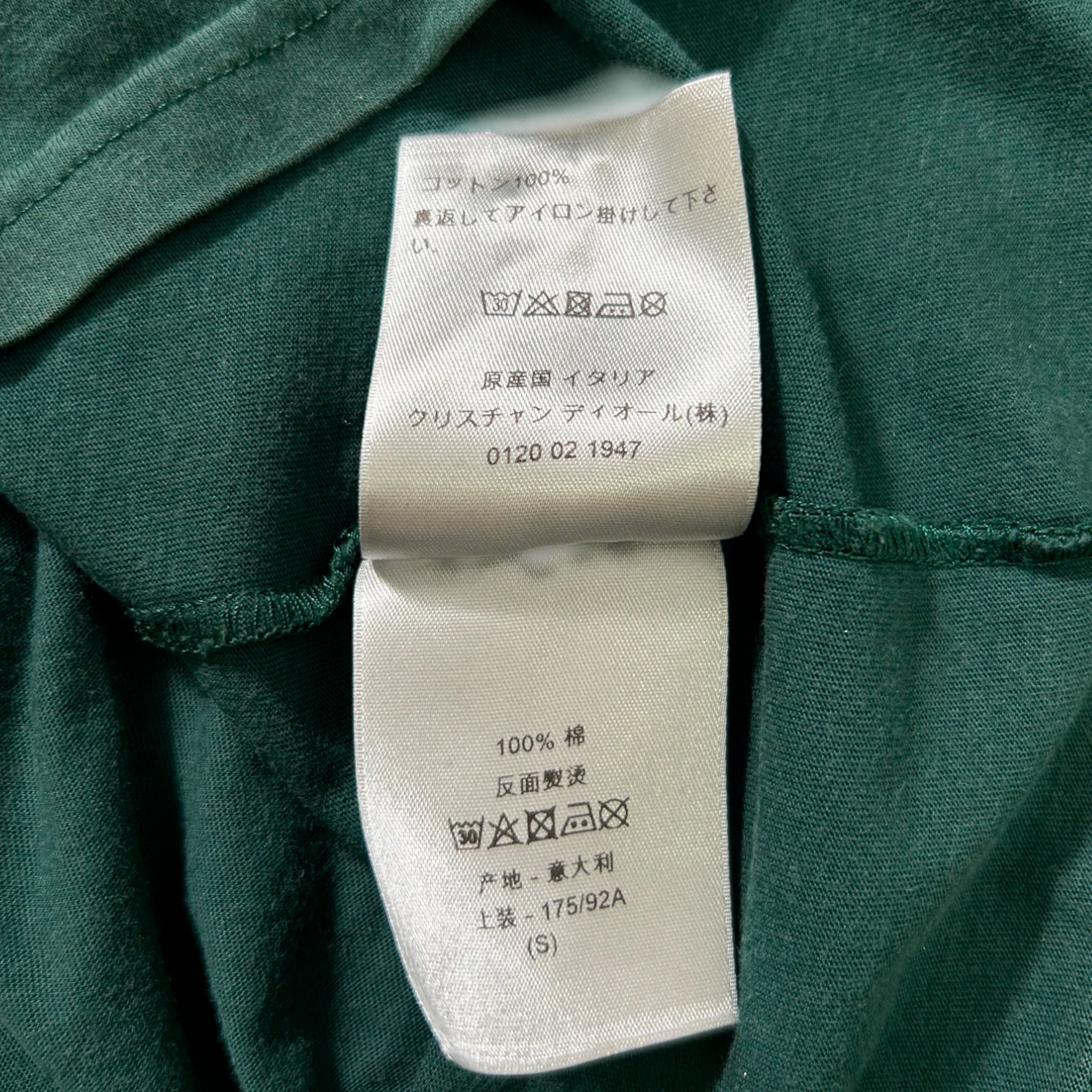 a close-up view of a green fabric, possibly a shirt or garment, with a white label attached to it. The label contains text in Japanese and English, providing information about the product.