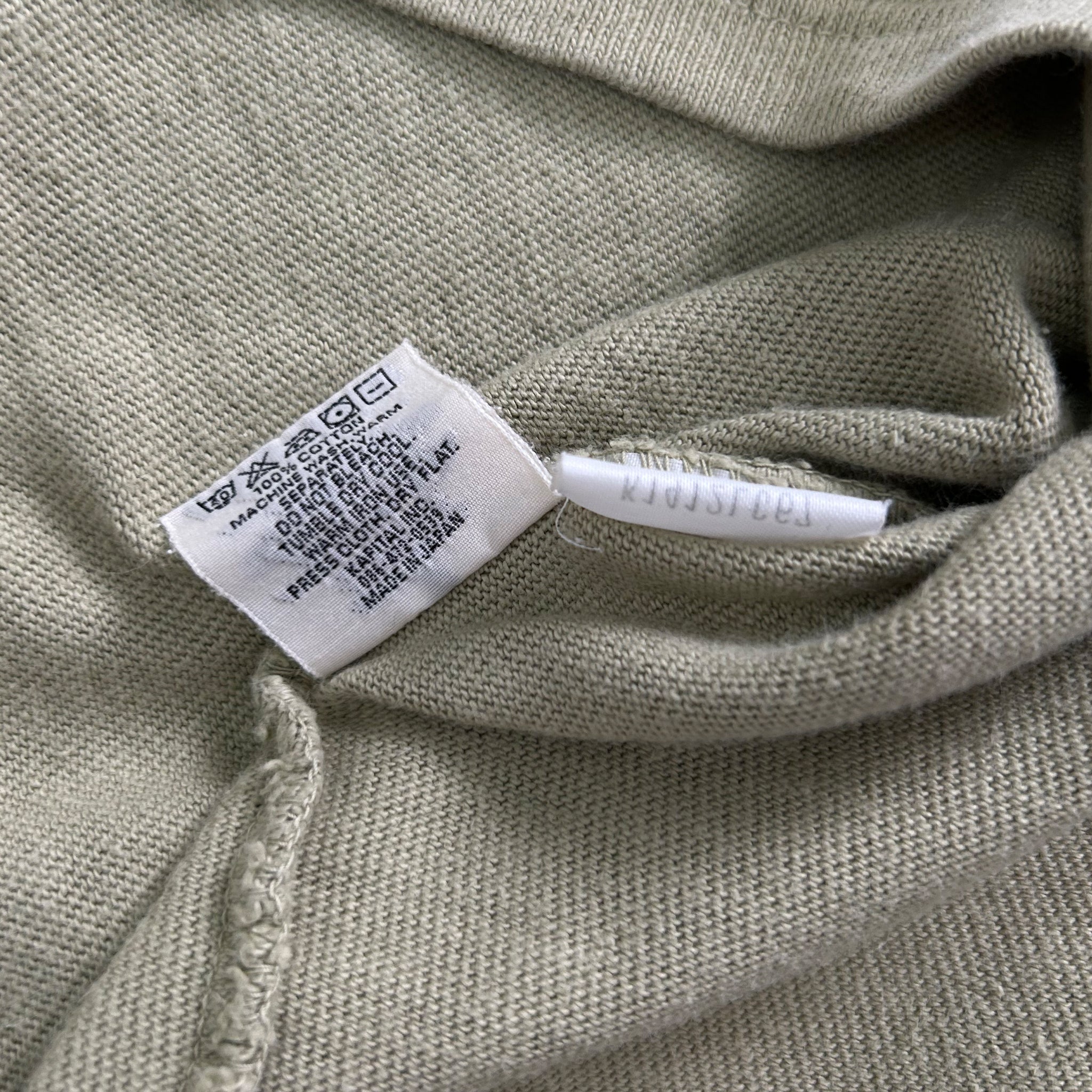 a close-up view of a grey fabric, possibly a sweater or a shirt, with a white label attached to it. The label appears to contain text and symbols, but the image does not contain any text that can be transcribed in less than 500 characters.