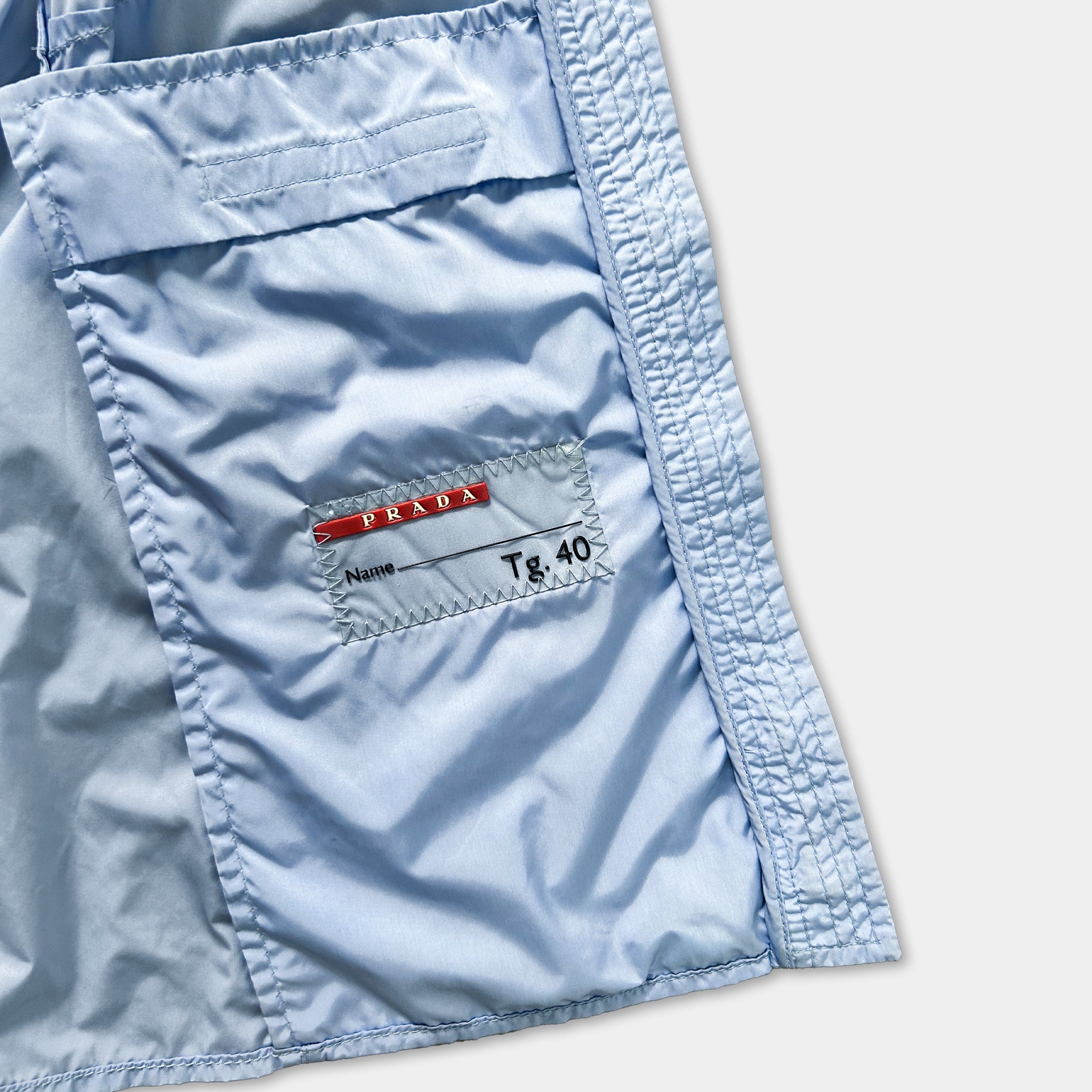 a close-up view of a light blue fabric, possibly a jacket or a shirt, with a small label or tag attached to it. The label appears to be a name tag or a label with a number, indicating the garment's brand or size.