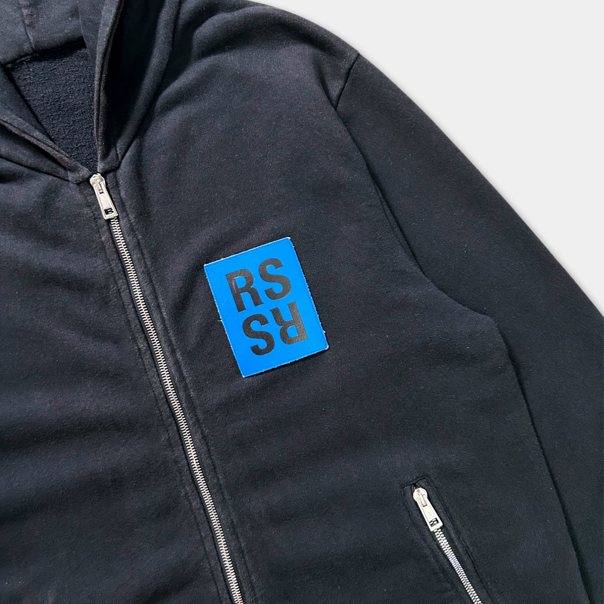 A black zip-up hoodie with a blue patch on the front.