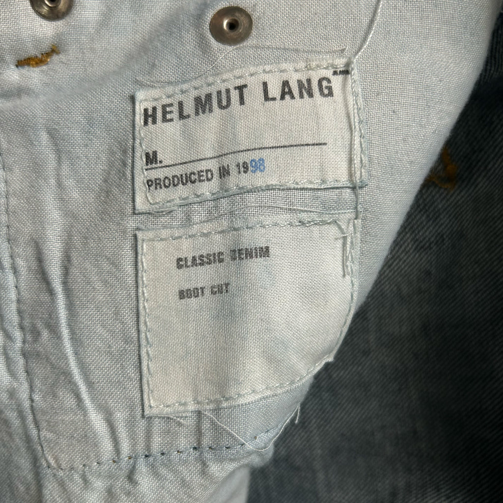 a close-up view of a denim fabric, possibly a piece of clothing, with a label attached to it. The label appears to be from the brand Helmut Lang, indicating that the fabric is produced in 1958 and is a classic denim boot cut.
