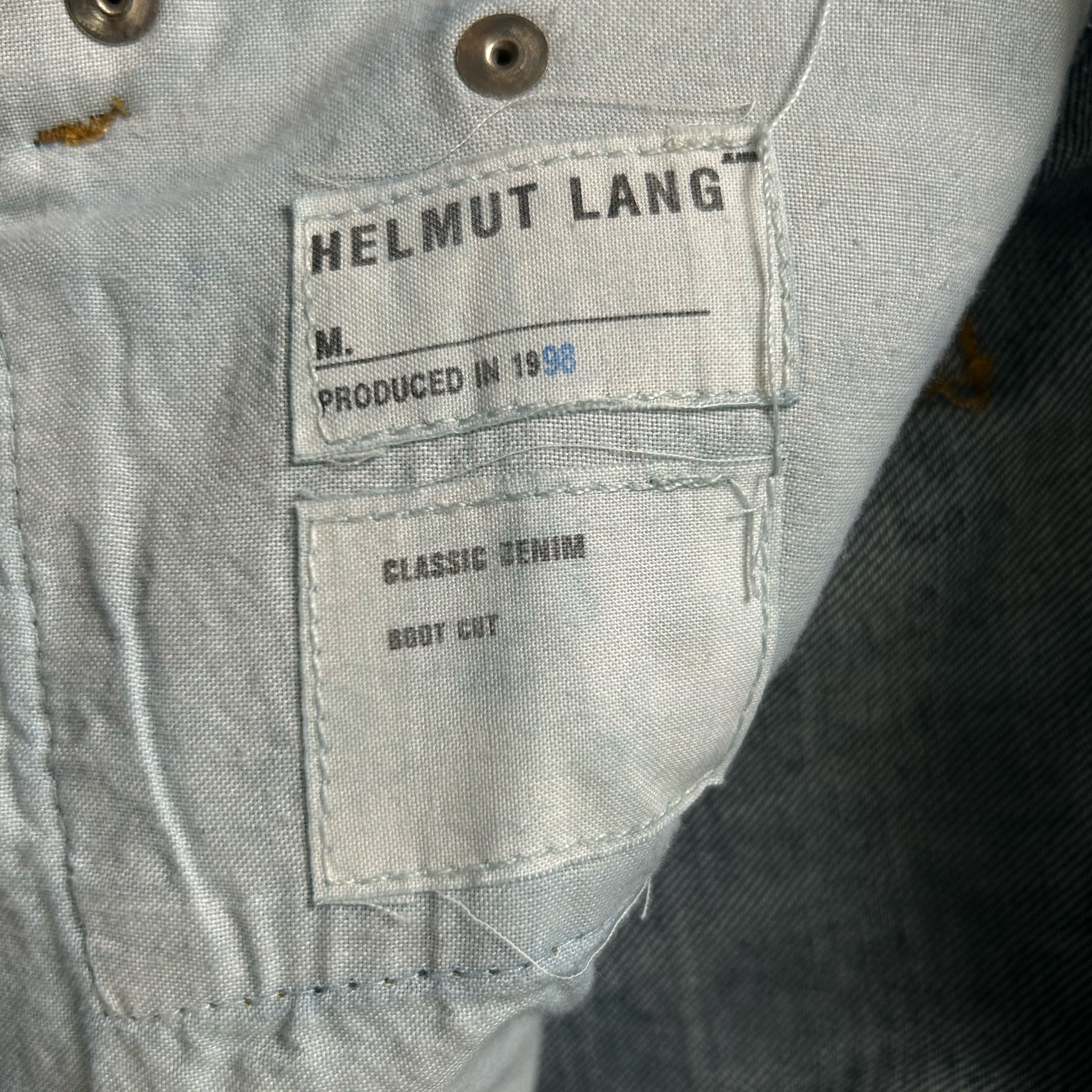 a close-up view of a denim fabric, possibly a piece of clothing, with a label attached to it. The label appears to be from the brand Helmut Lang, indicating that the fabric is produced in 1958 and is a classic denim boot cut.