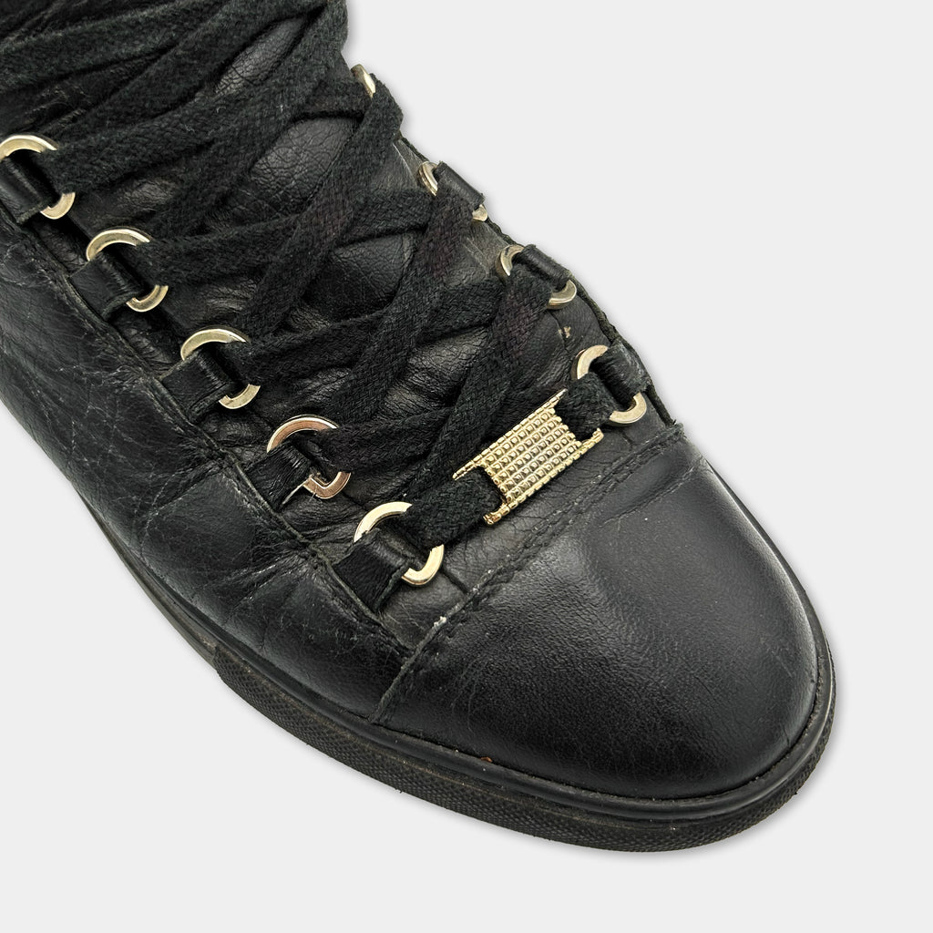 A close-up view of a black leather sneaker with a gold-colored metal buckle on the side.