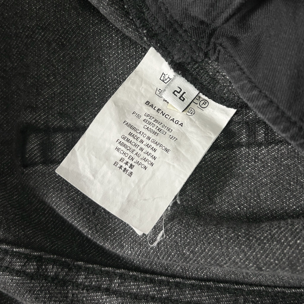 a close-up view of a black fabric, possibly denim, with a white label attached to it. The label appears to be a garment tag or label from a clothing item, possibly a pair of jeans. The label contains text in a foreign language, which suggests that the image may be from a different country or region. The text on the label is not very clear, and it is not possible to transcribe it accurately without more context or a clearer view of the label.